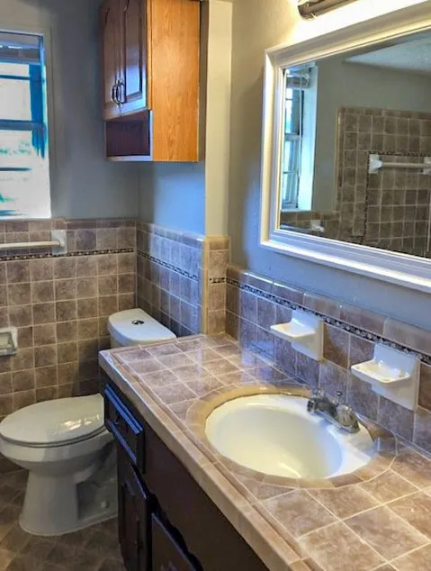 Bathroom featuring a tile-top vanity with an integrated sink, a dark wood-finish cabinet, and a framed wall mirror with overhead lighting