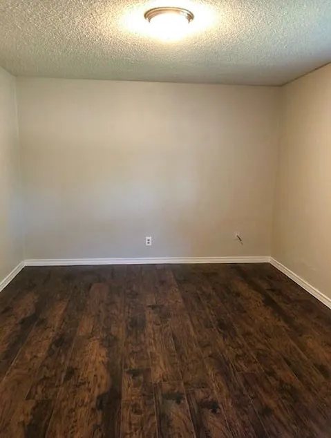 Spacious room featuring rich wood-finish flooring, light neutral walls, white baseboards, and a flush-mount ceiling light fixture