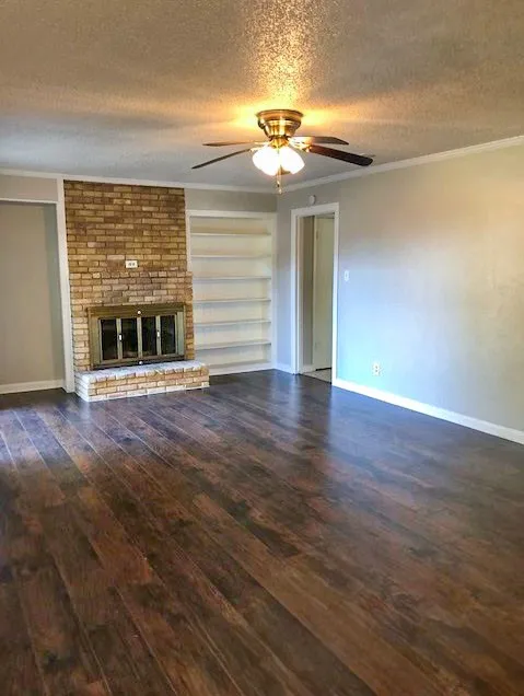 Spacious room featuring rich wood-finish flooring, a brick fireplace with a metal insert, a ceiling fan with integrated lighting, built-in shelving, and light-toned walls