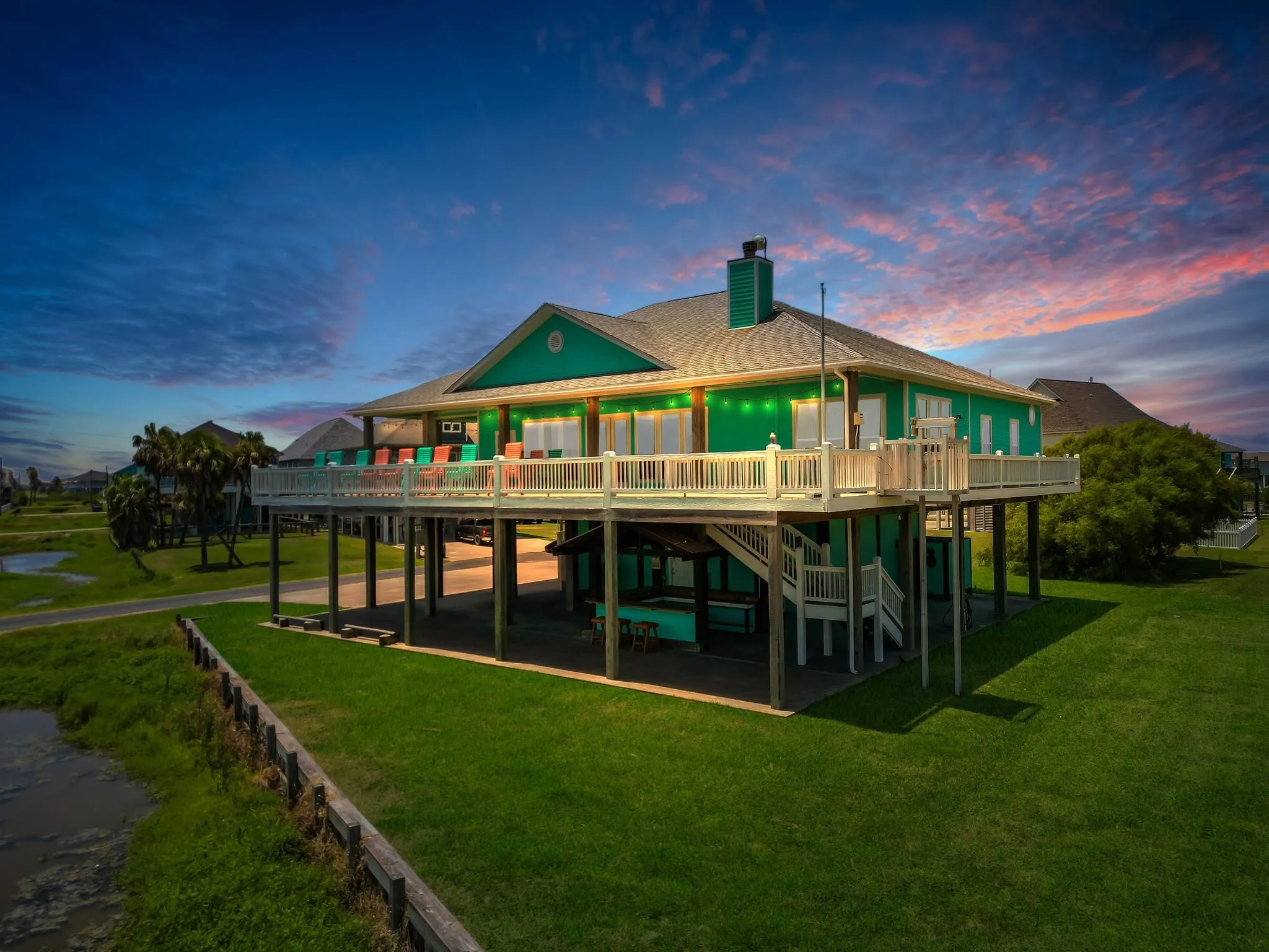 Elevated Coastal Contemporary home featuring a striking green metal roof, expansive wraparound deck with white railings, and open-air covered carport beneath