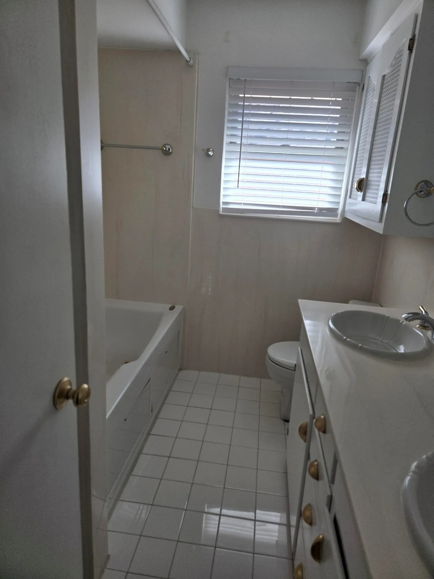 White tile flooring, a built-in bathtub, a window with horizontal blinds, a toilet, and a vanity with two integrated sinks