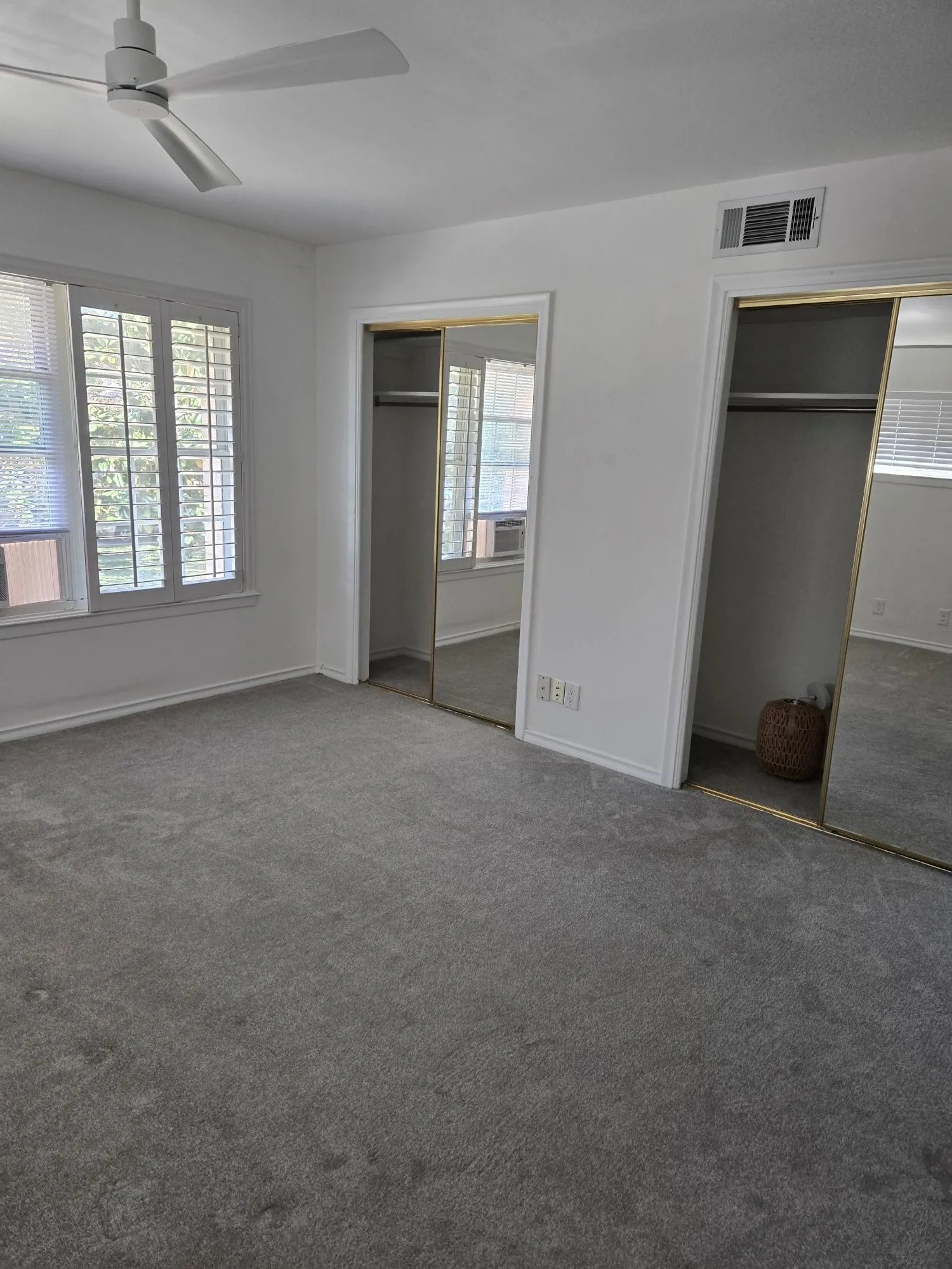 Ceiling fan, window with white plantation shutters, two mirrored closet doors, and grey carpet flooring