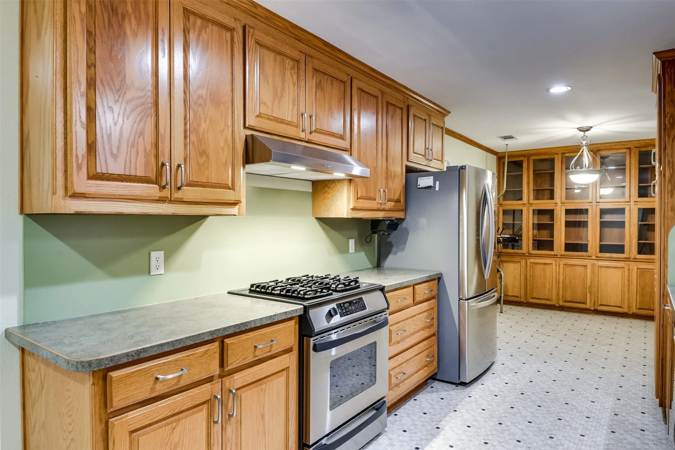 Kitchen featuring appliances with stainless steel finishes, light floors, under cabinet range hood, decorative light fixtures, and brown cabinets