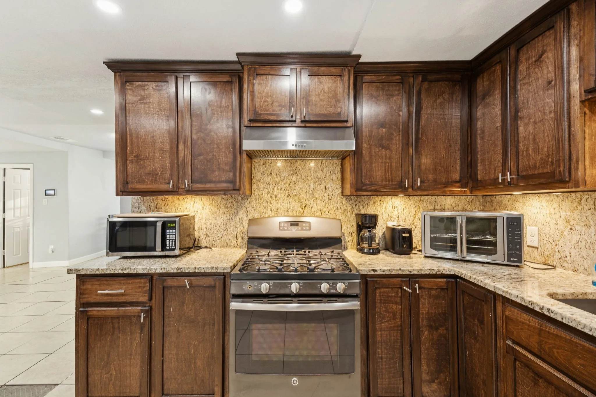Kitchen with stainless steel appliances, backsplash, light stone counters, range hood, and dark brown cabinetry