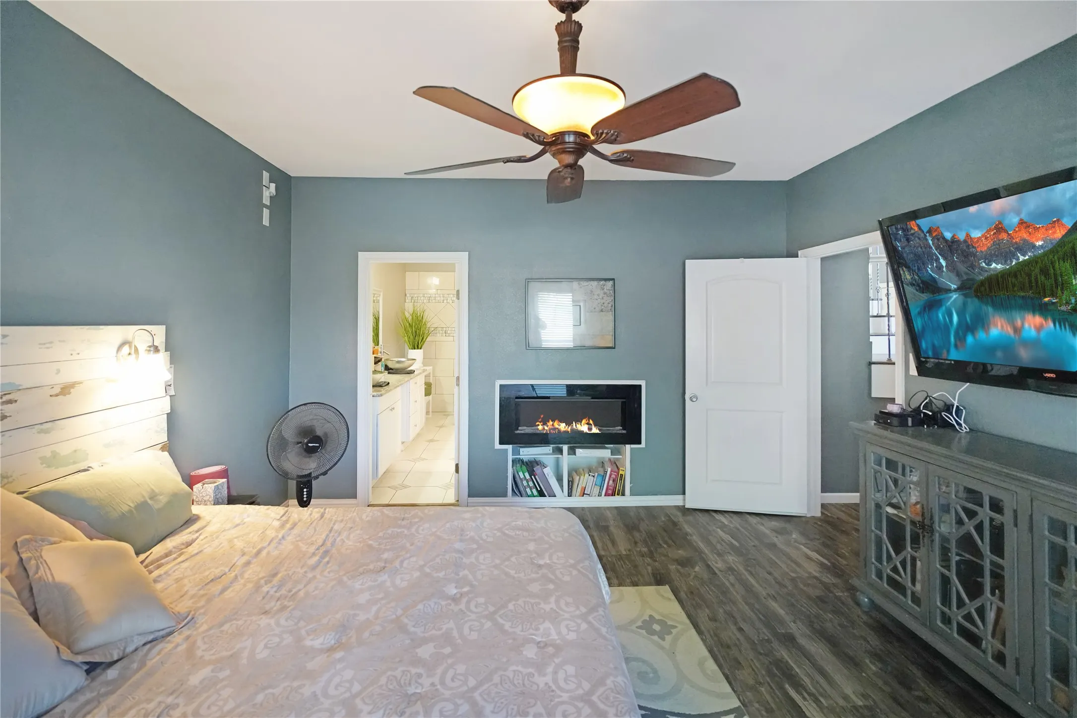 Bedroom with dark wood-style flooring, ceiling fan, a glass covered fireplace, and ensuite bathroom
