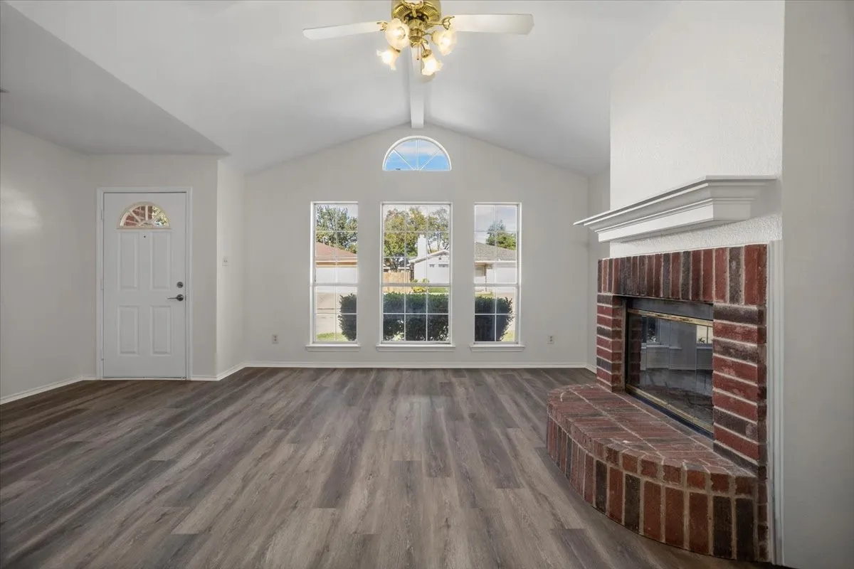 Unfurnished living room featuring a brick fireplace, dark wood-style flooring, vaulted ceiling, and a ceiling fan