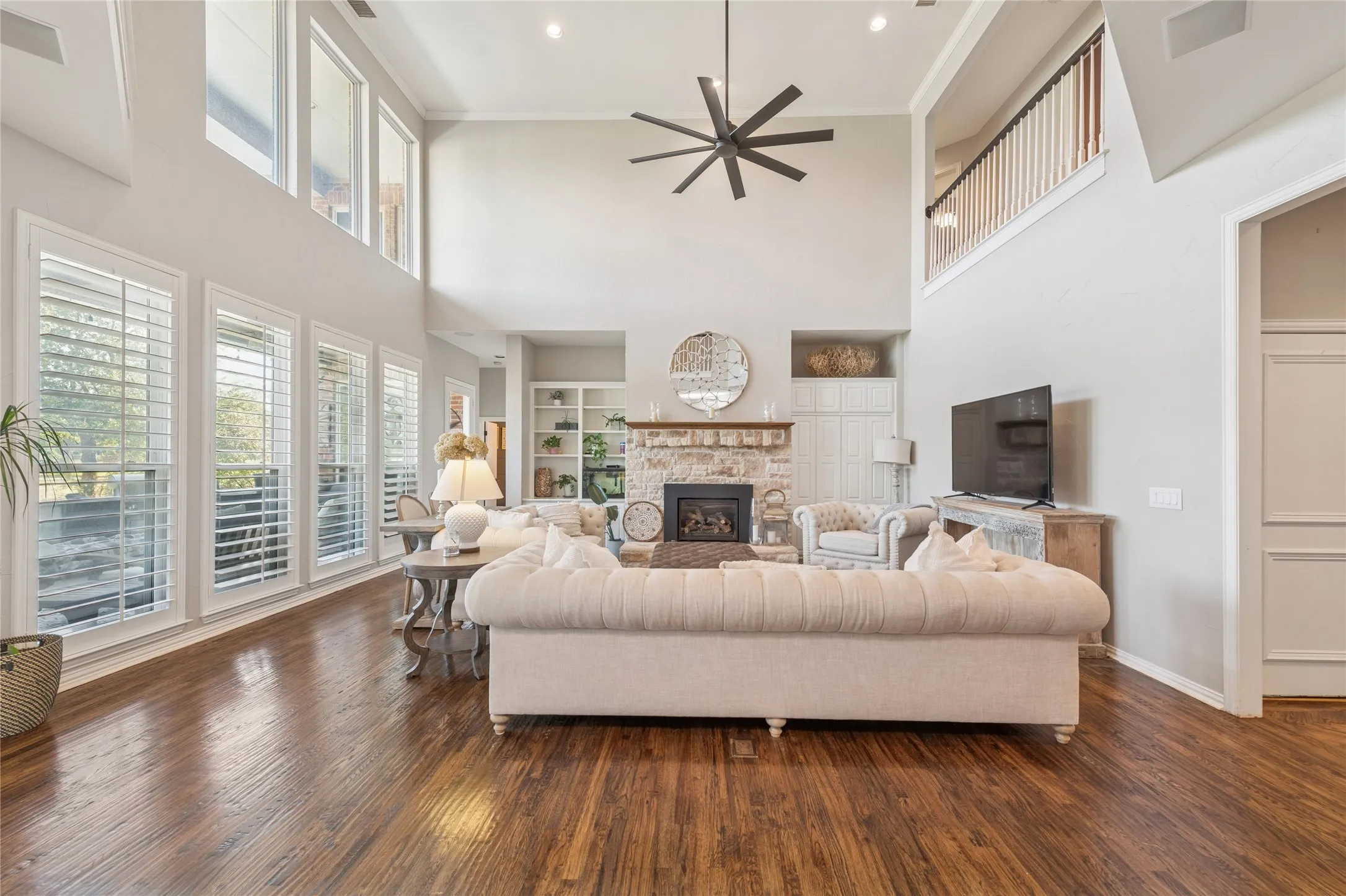 Living room with a stone fireplace, built in features, ceiling fan, dark wood-style flooring, and a towering ceiling