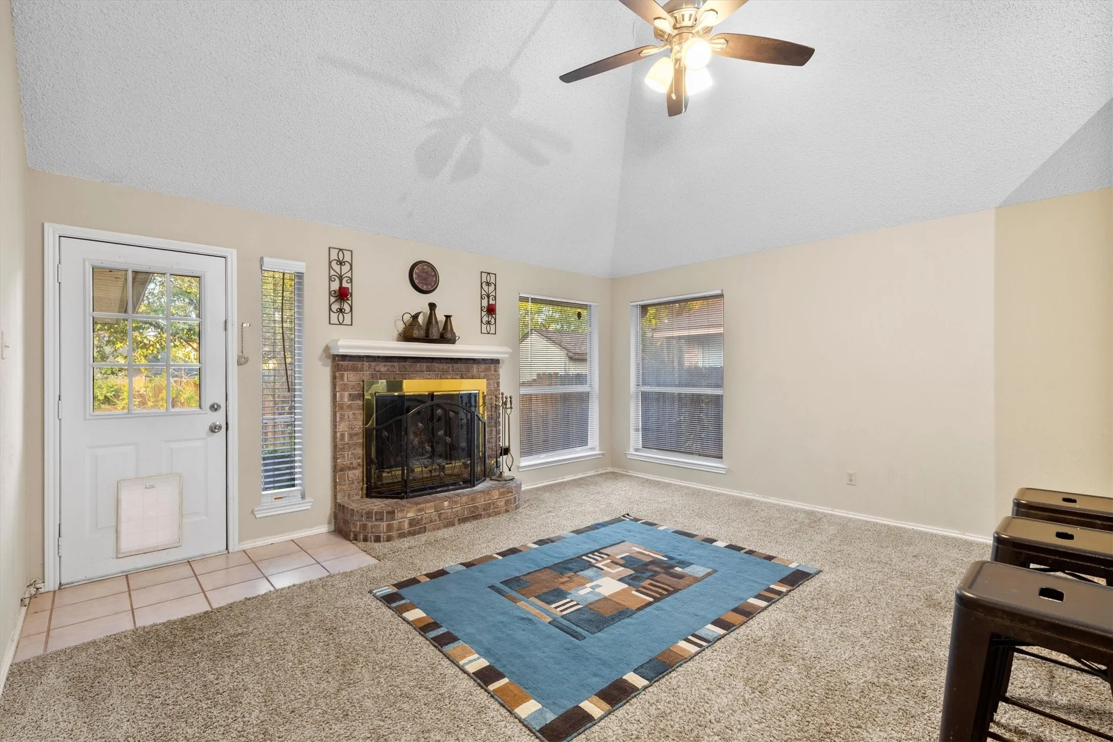 Carpeted living area with vaulted ceiling, a brick fireplace, a textured ceiling, a ceiling fan, and tile patterned floors