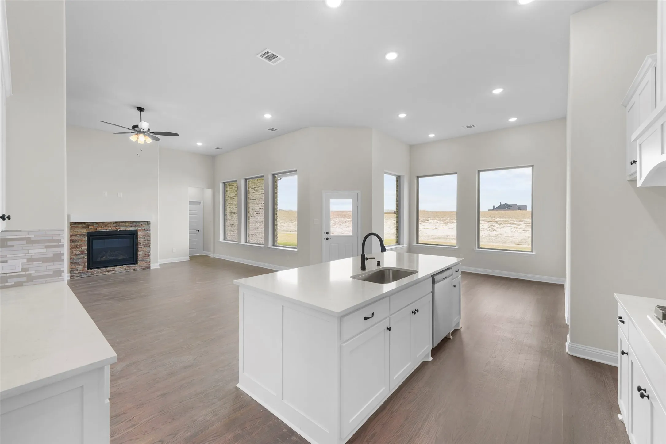 Open-concept living space featuring wood-finish flooring, a kitchen island with an undermount sink, white cabinetry, a stone-clad fireplace, and recessed lighting