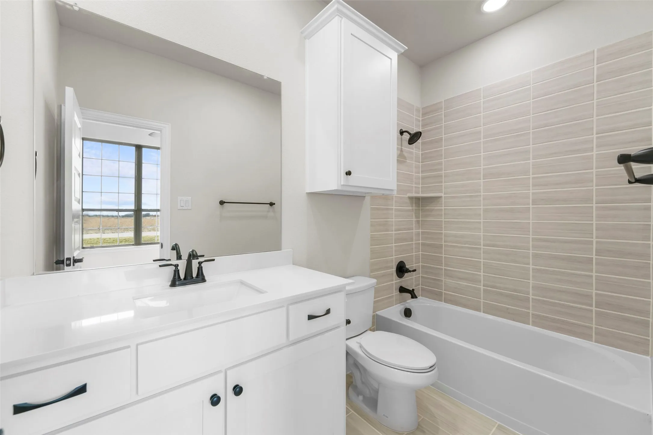White vanity with an integrated sink and matte black faucet, full-width mirror, built-in white cabinetry, bathtub with subway tile surround, and light-toned tile flooring