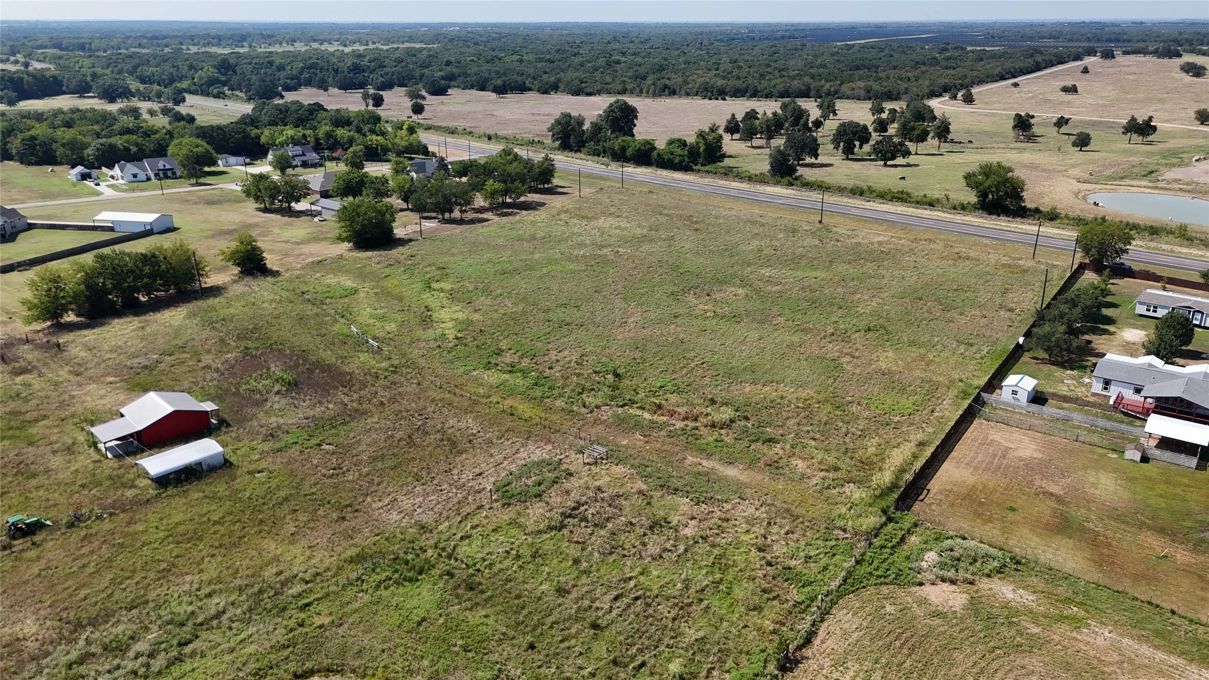 Expansive rural landscape featuring a cleared parcel of land, a visible roadway, scattered mature trees, and a small pond