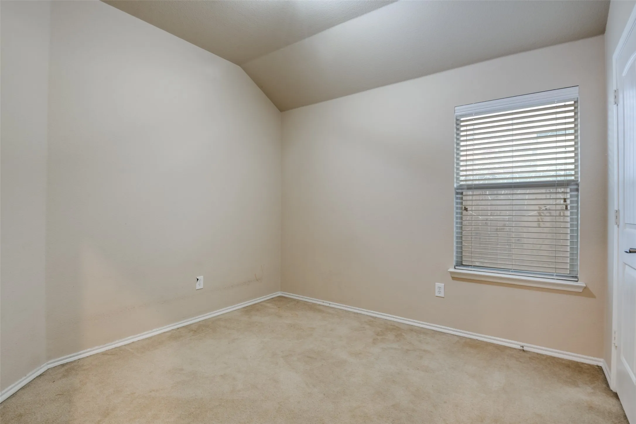 Carpeted room featuring a single window with horizontal blinds, neutral wall paint, white baseboards, and a vaulted ceiling