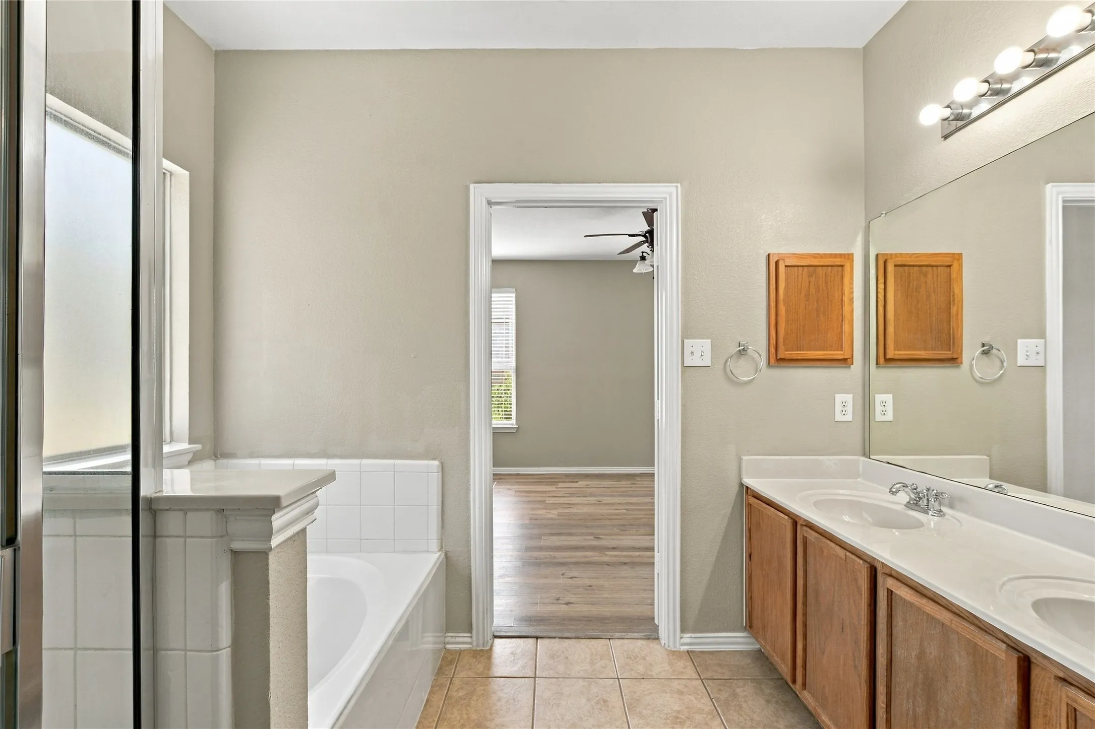 Double vanity with integrated sinks and chrome fixtures, large mirror with strip lighting, tiled flooring, and a built-in bathtub with tiled surround