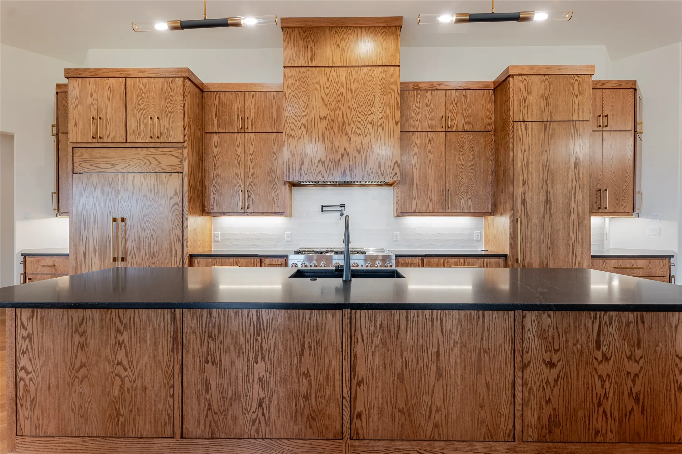 Kitchen with decorative backsplash, a large island, brown cabinets, stainless steel gas stovetop, and dark stone counters