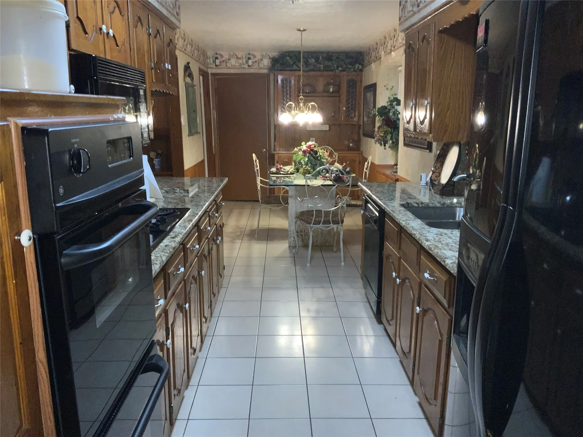 Galley kitchen featuring wood cabinetry, granite-finish countertops, black appliances, and tile flooring