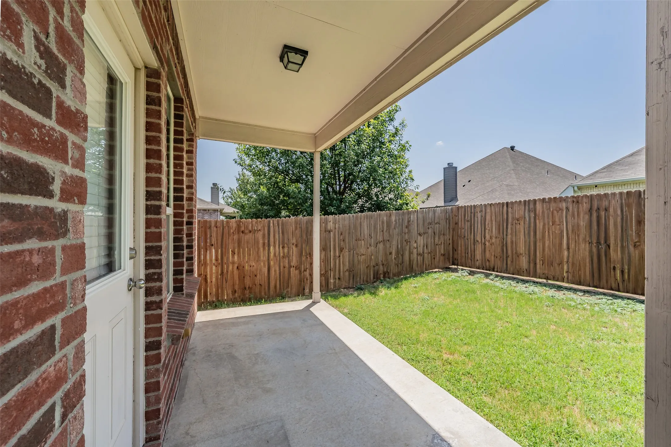 Covered patio with a concrete slab, red brick exterior, solid privacy fence, green lawn, and exterior lighting fixture