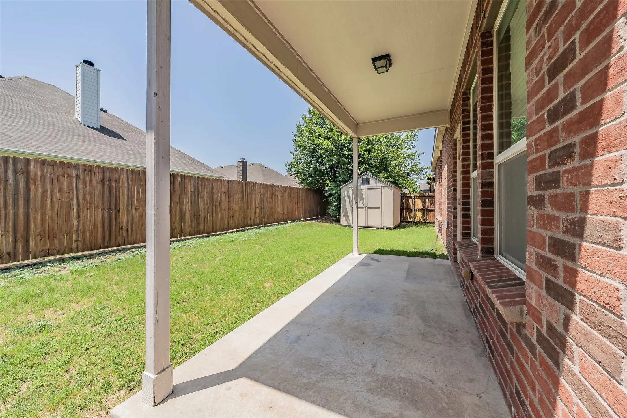 Covered concrete patio overlooking a private backyard with a wood privacy fence, green lawn, and an included storage shed
