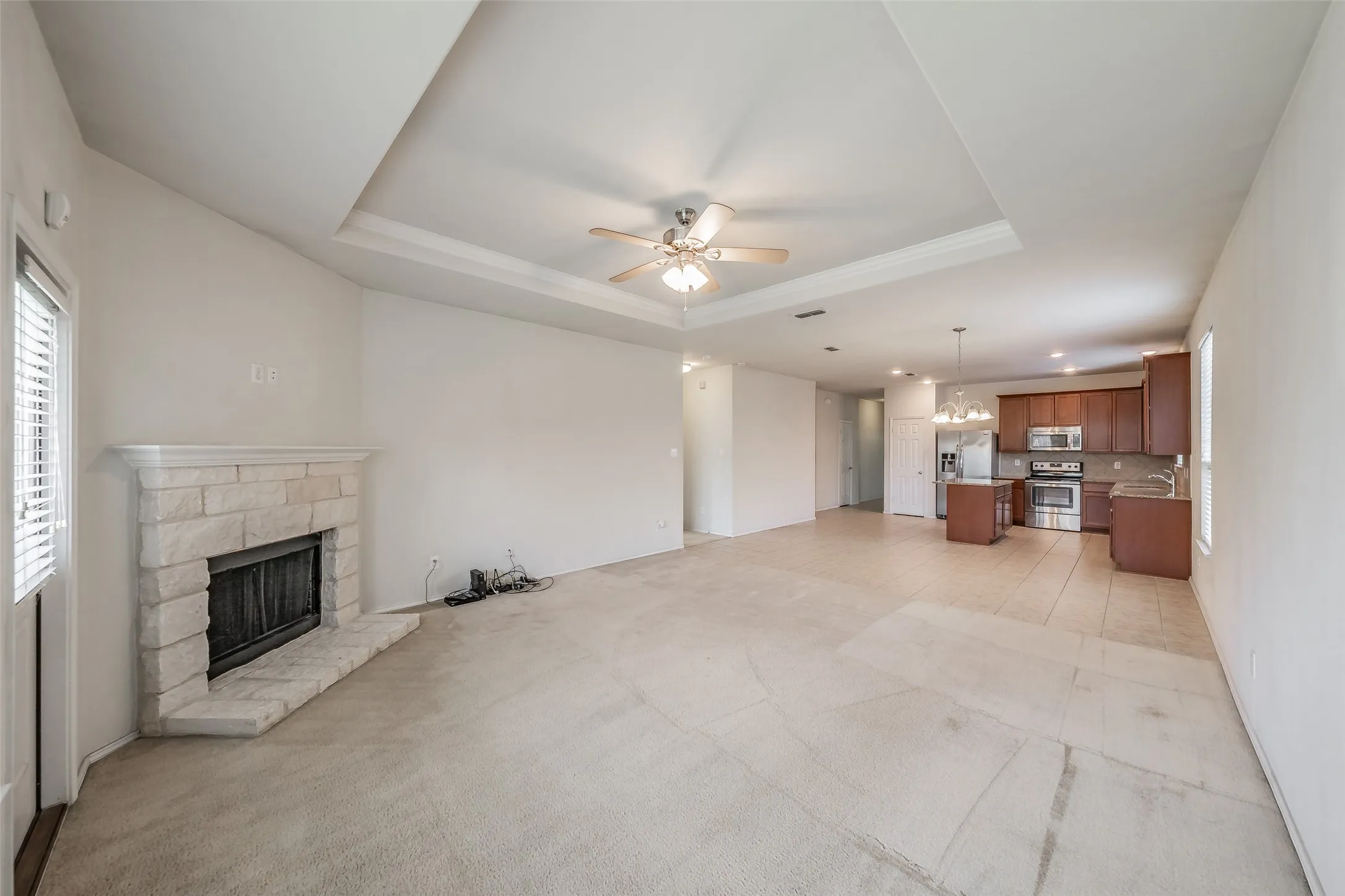 Spacious open-concept living area featuring a stone fireplace with hearth, a tray ceiling with a ceiling fan, and light-toned carpet