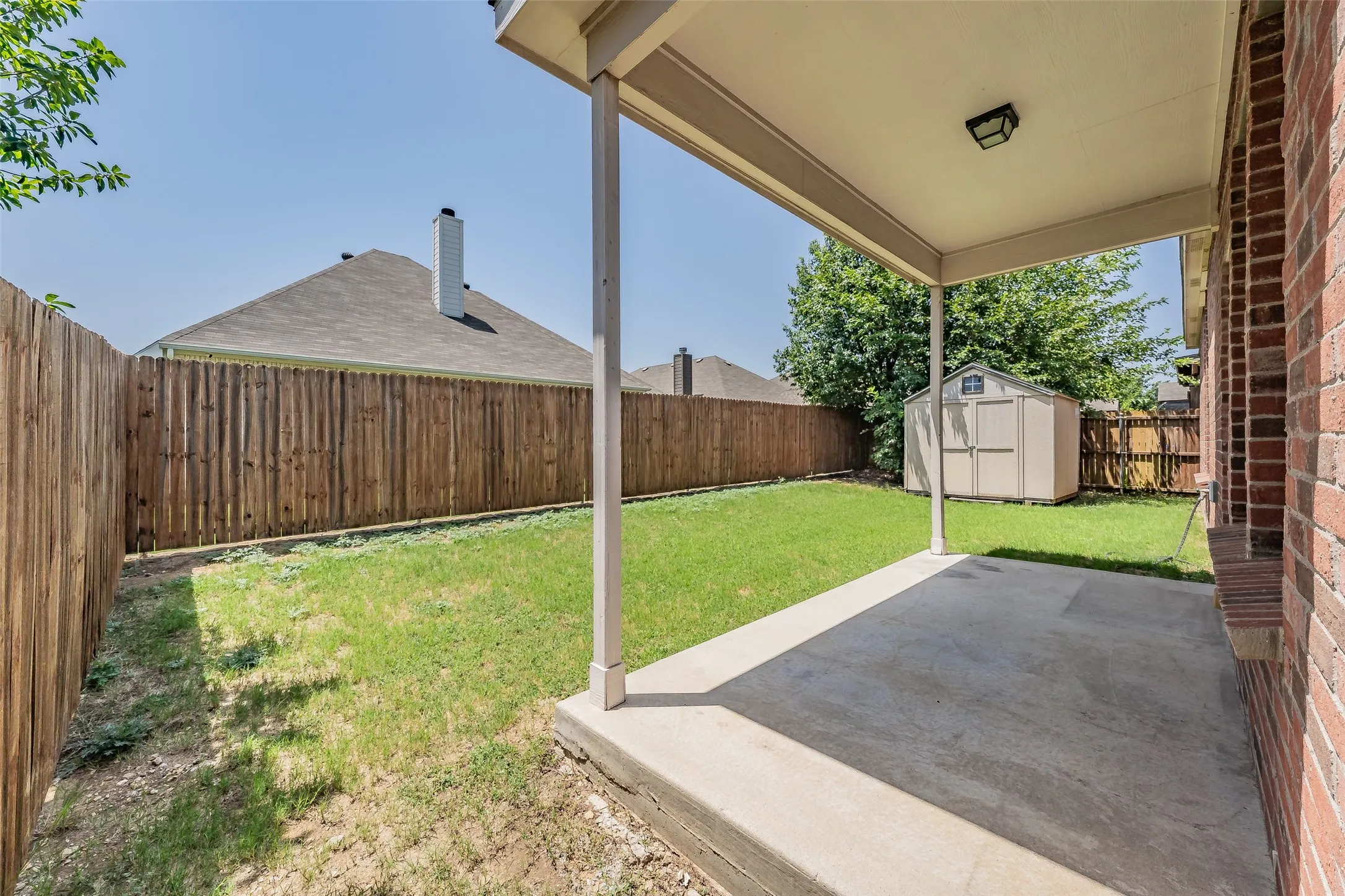 Covered concrete patio with a brick exterior, offering direct access to a fenced backyard featuring a storage shed and mature landscaping