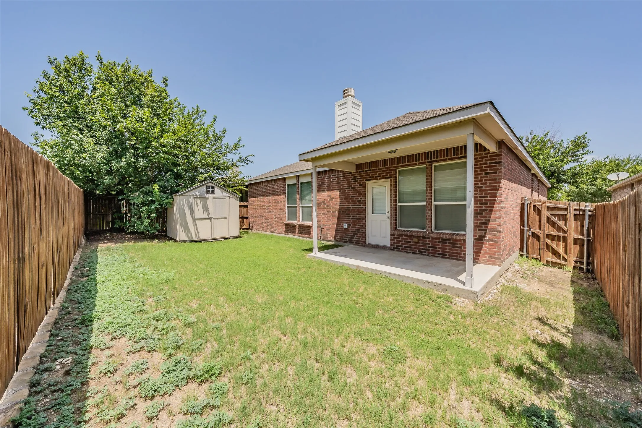 Enclosed backyard featuring a brick exterior, covered patio, wood fencing, and a storage shed