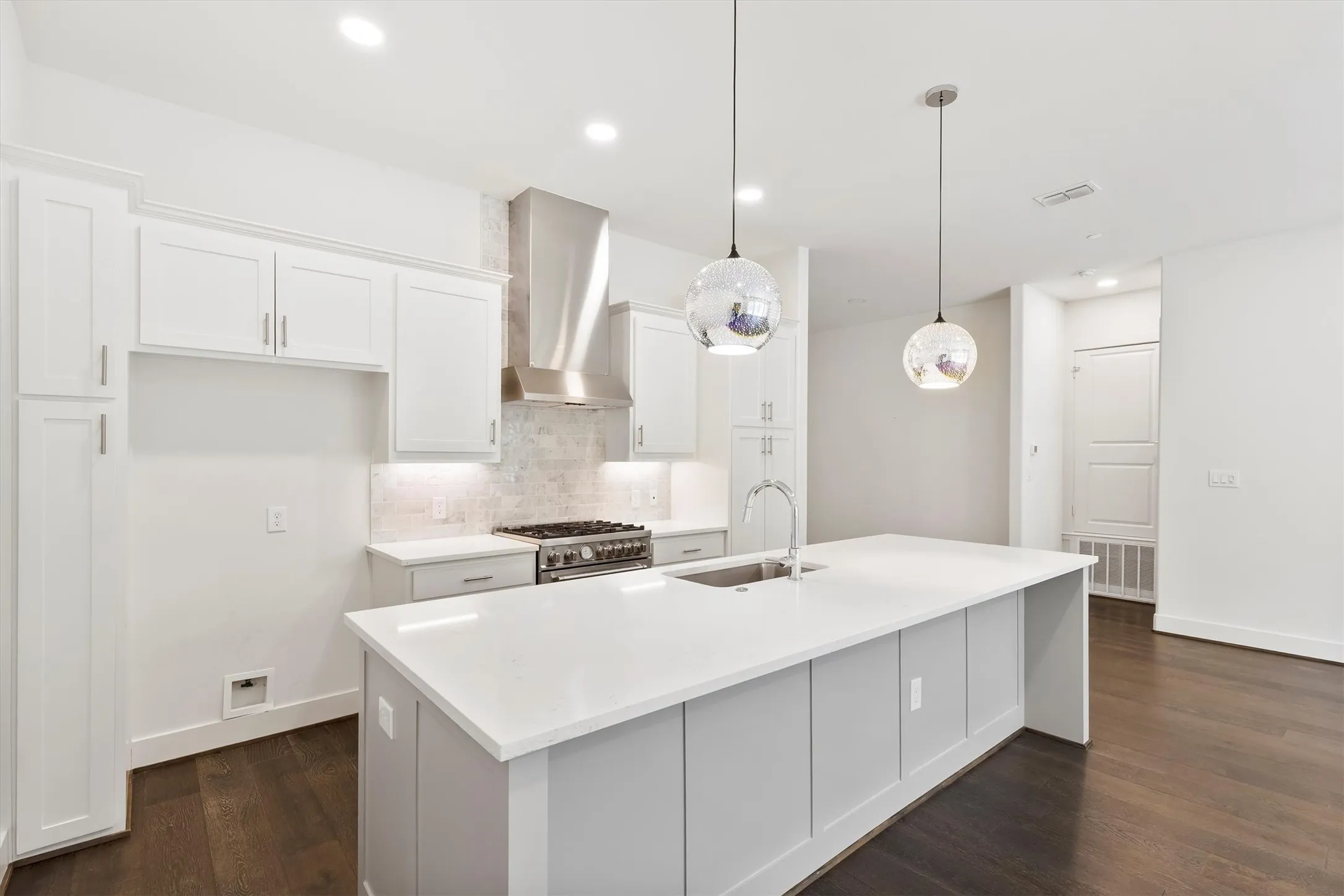 Kitchen featuring white cabinets, wall chimney range hood, backsplash, hanging light fixtures, and a center island with sink