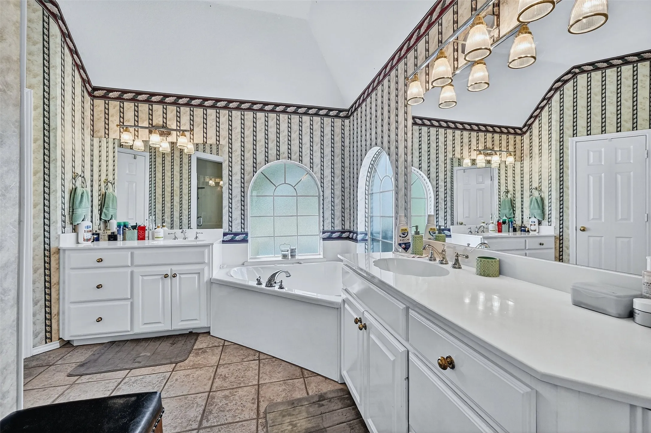 Full bath with lofted ceiling, a chandelier, wallpapered walls, a bath, and two vanities
