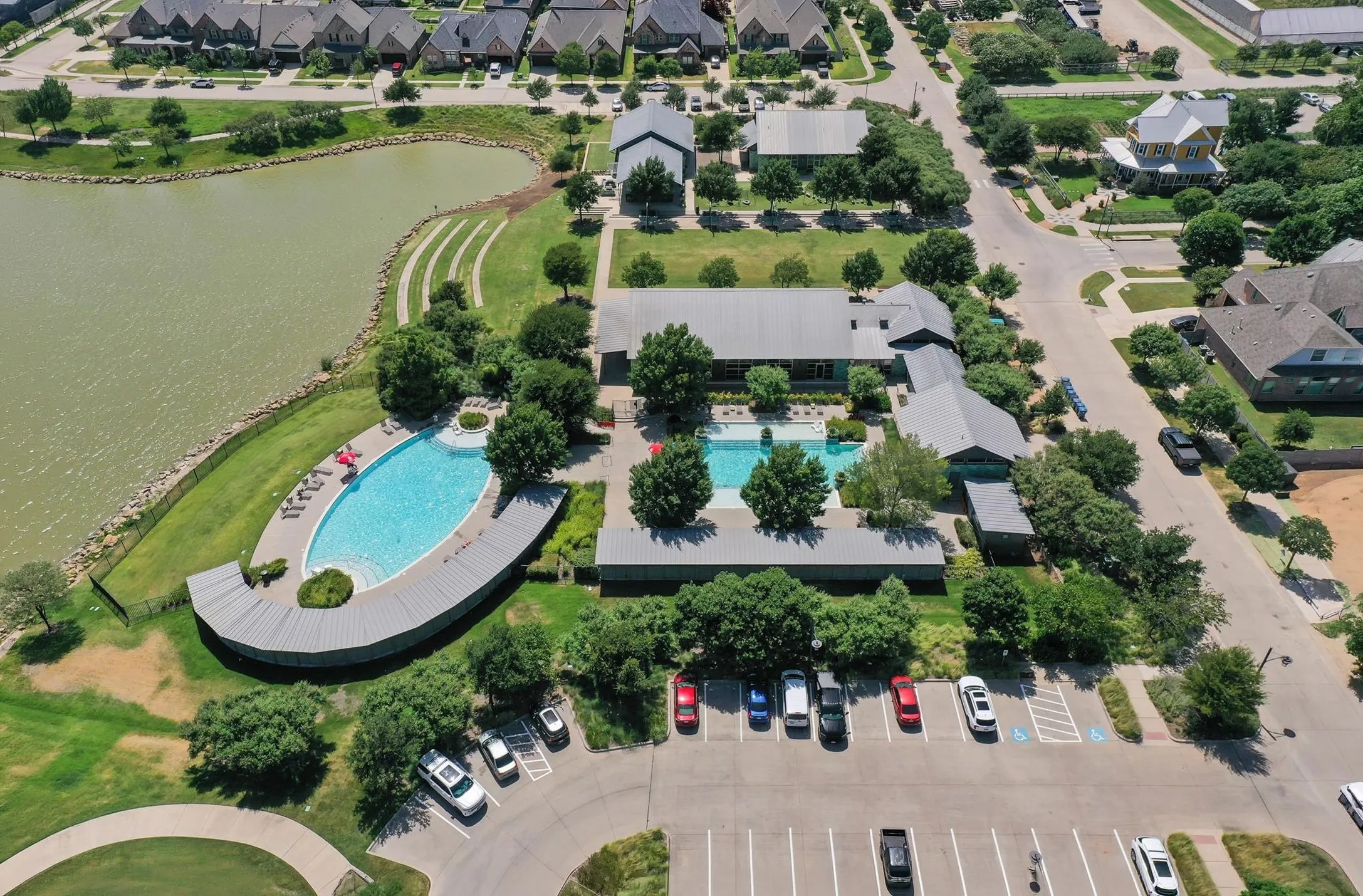 Aerial view of a community featuring two swimming pools, a large body of water, extensive green spaces, and dedicated parking areas