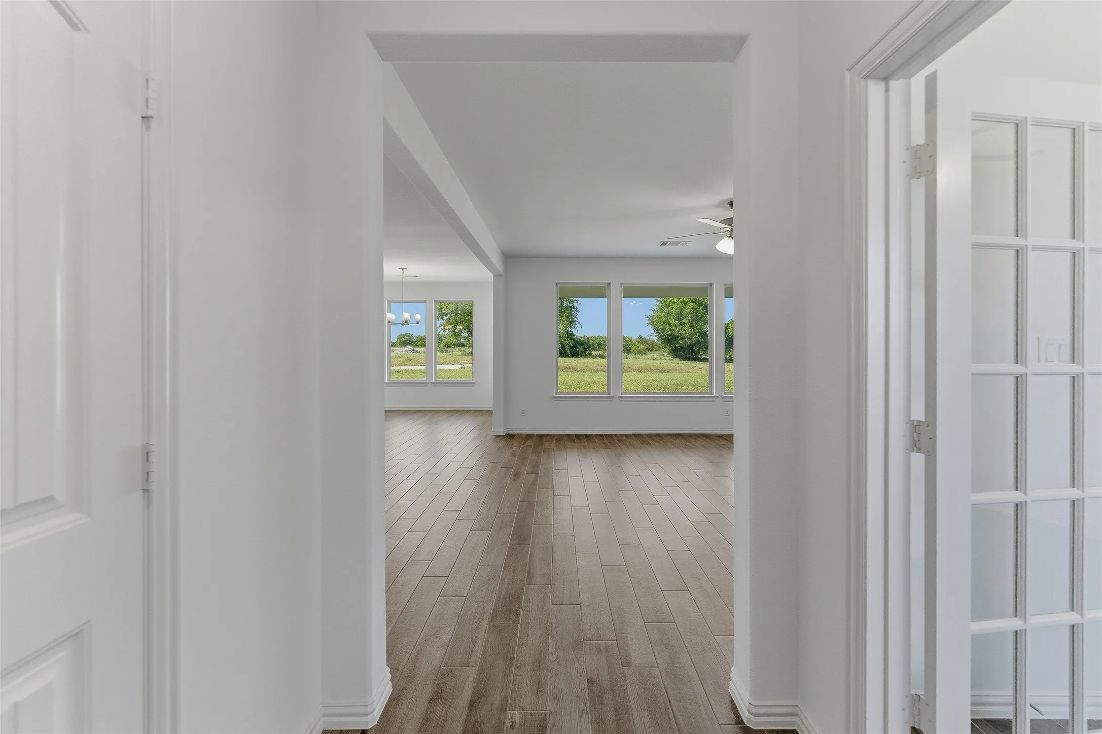 Hallway with wood-finish flooring, leading into an open living area featuring large windows, a ceiling fan, and a chandelier