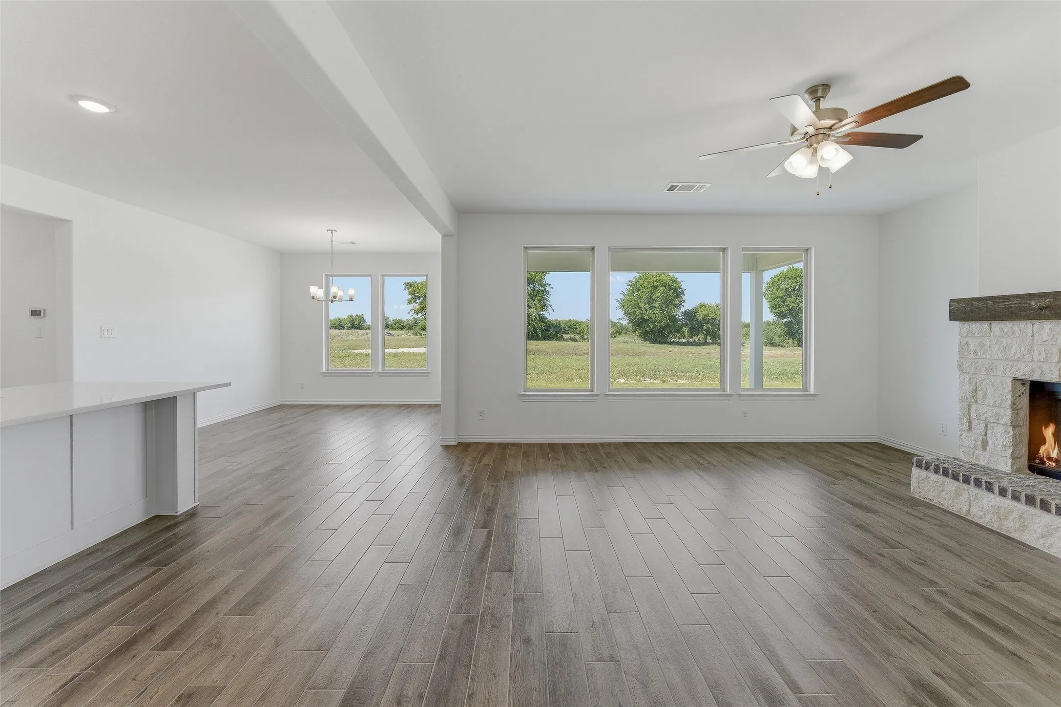 Open-concept living space featuring wood-finish flooring, a stone fireplace with a substantial mantle, and multiple large windows overlooking exterior scenery