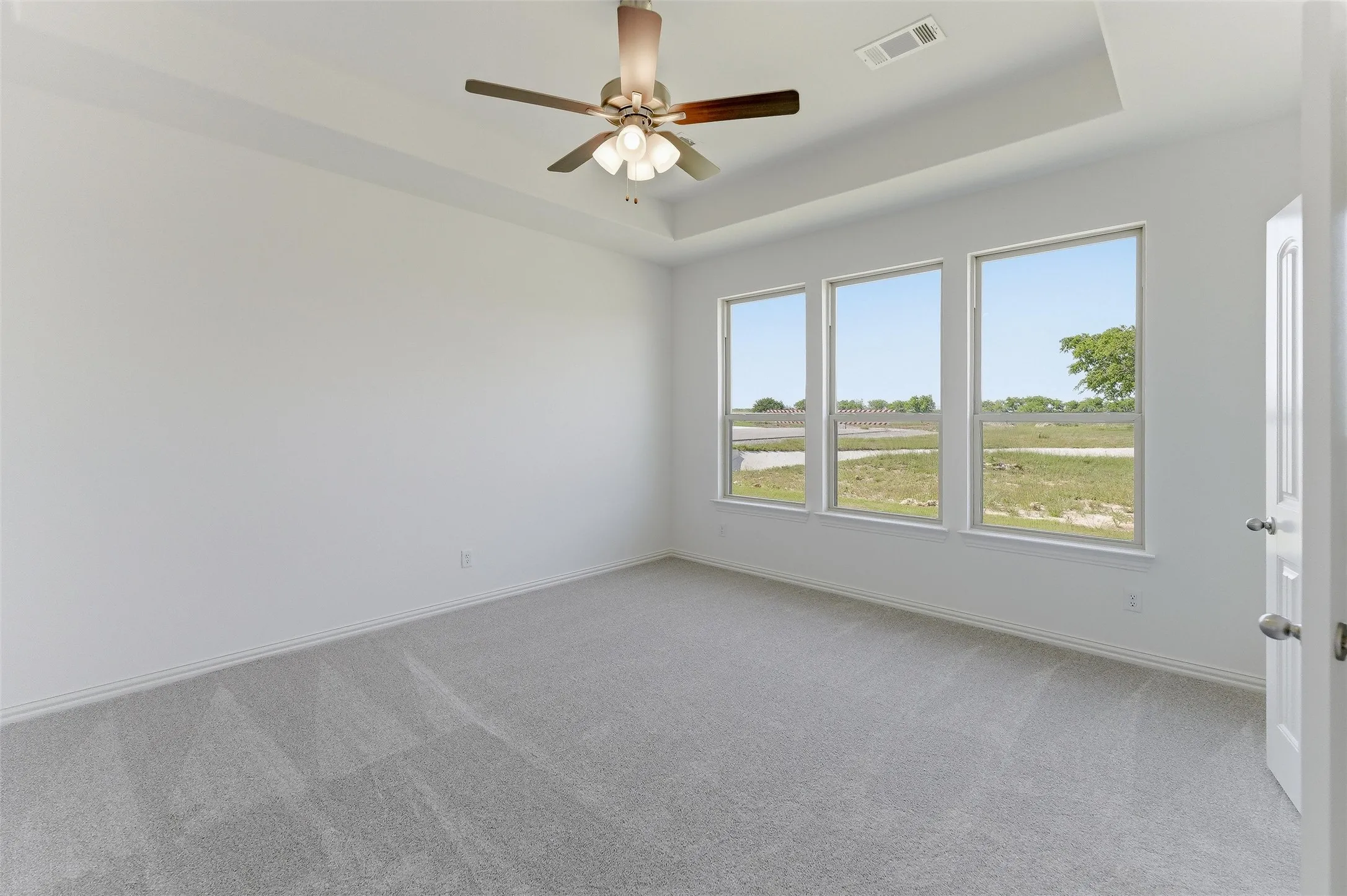 Spacious room featuring three large windows, light gray carpeting, and a white ceiling fan with integrated lighting