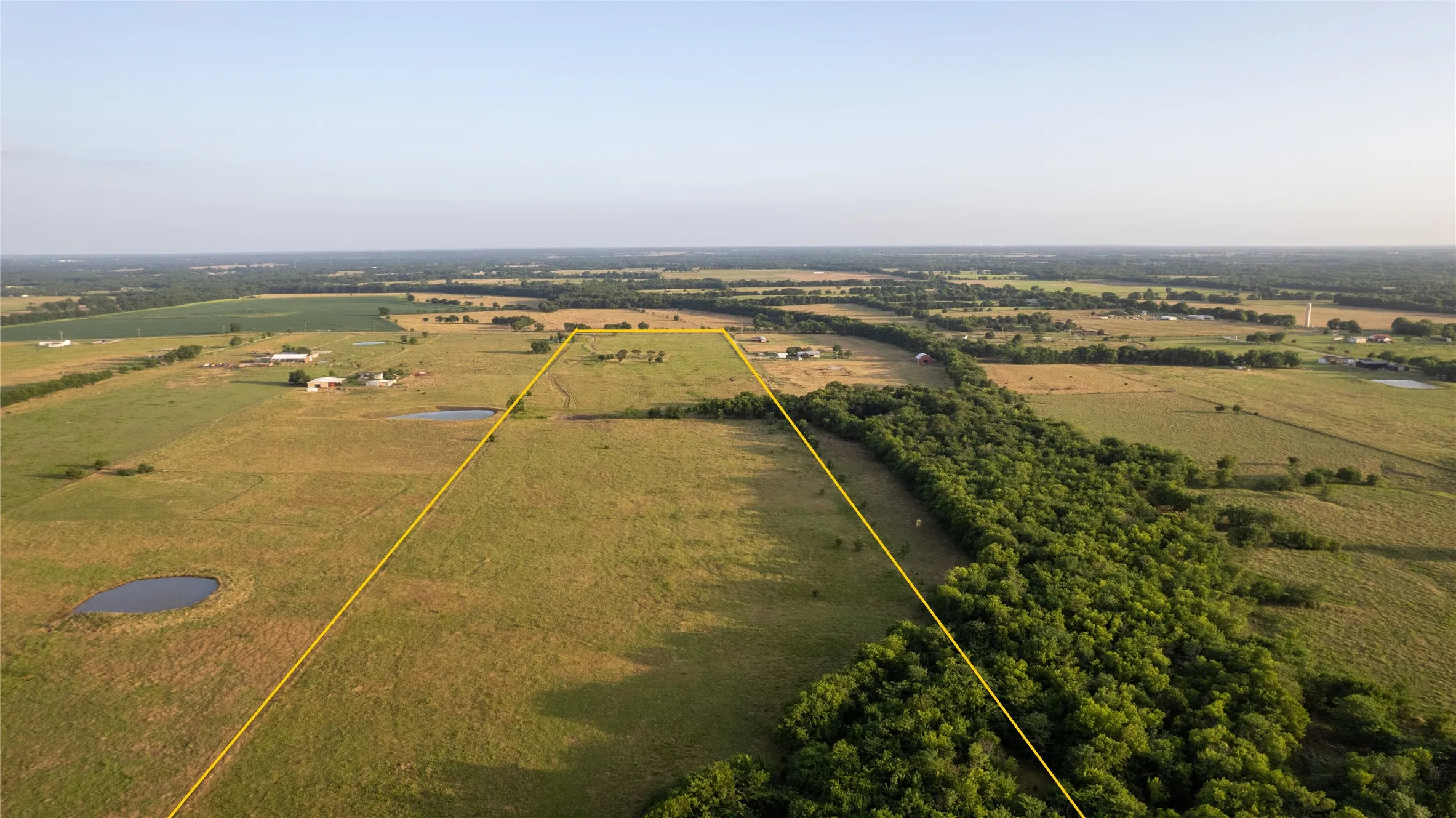 Expansive agricultural land featuring a natural pond, a dense tree line, and open fields