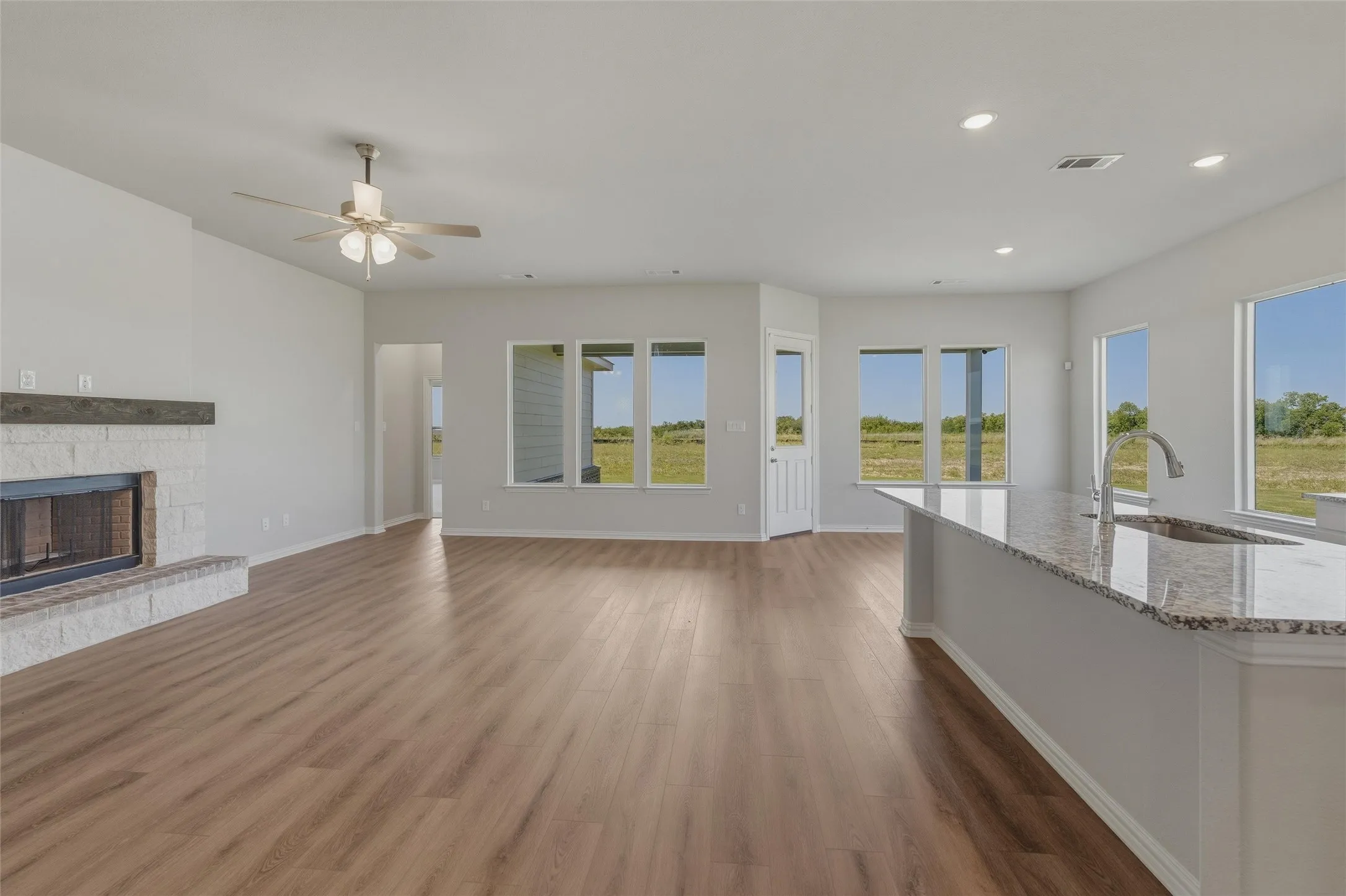 Open-concept living area featuring wood-finish flooring, a white brick fireplace with a wood mantle, and an integrated kitchen island with a granite-style countertop and undermount sink