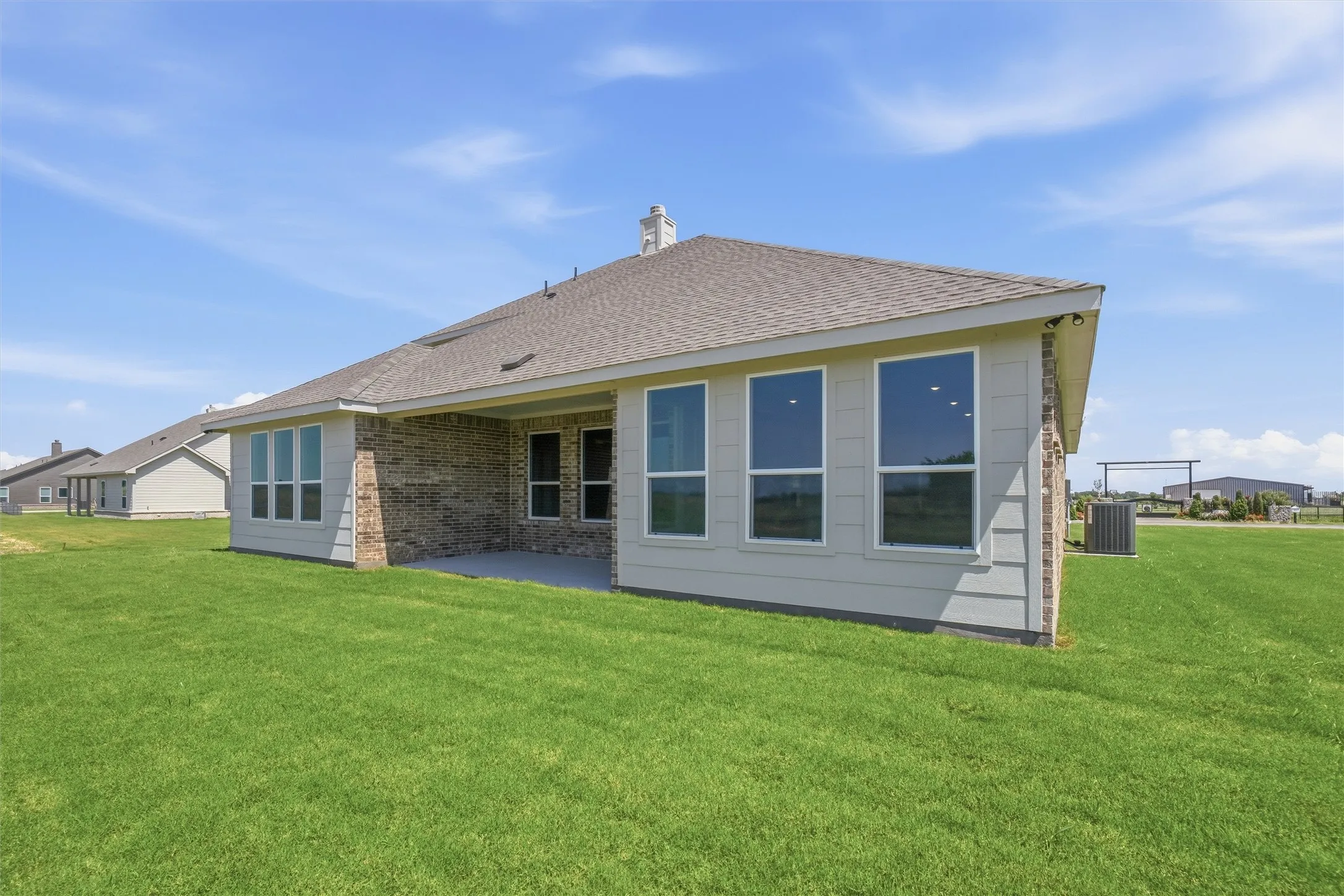 Expansive green lawn surrounds the property, featuring a covered patio, brick and siding exterior, multiple large windows, and a shingled roof
