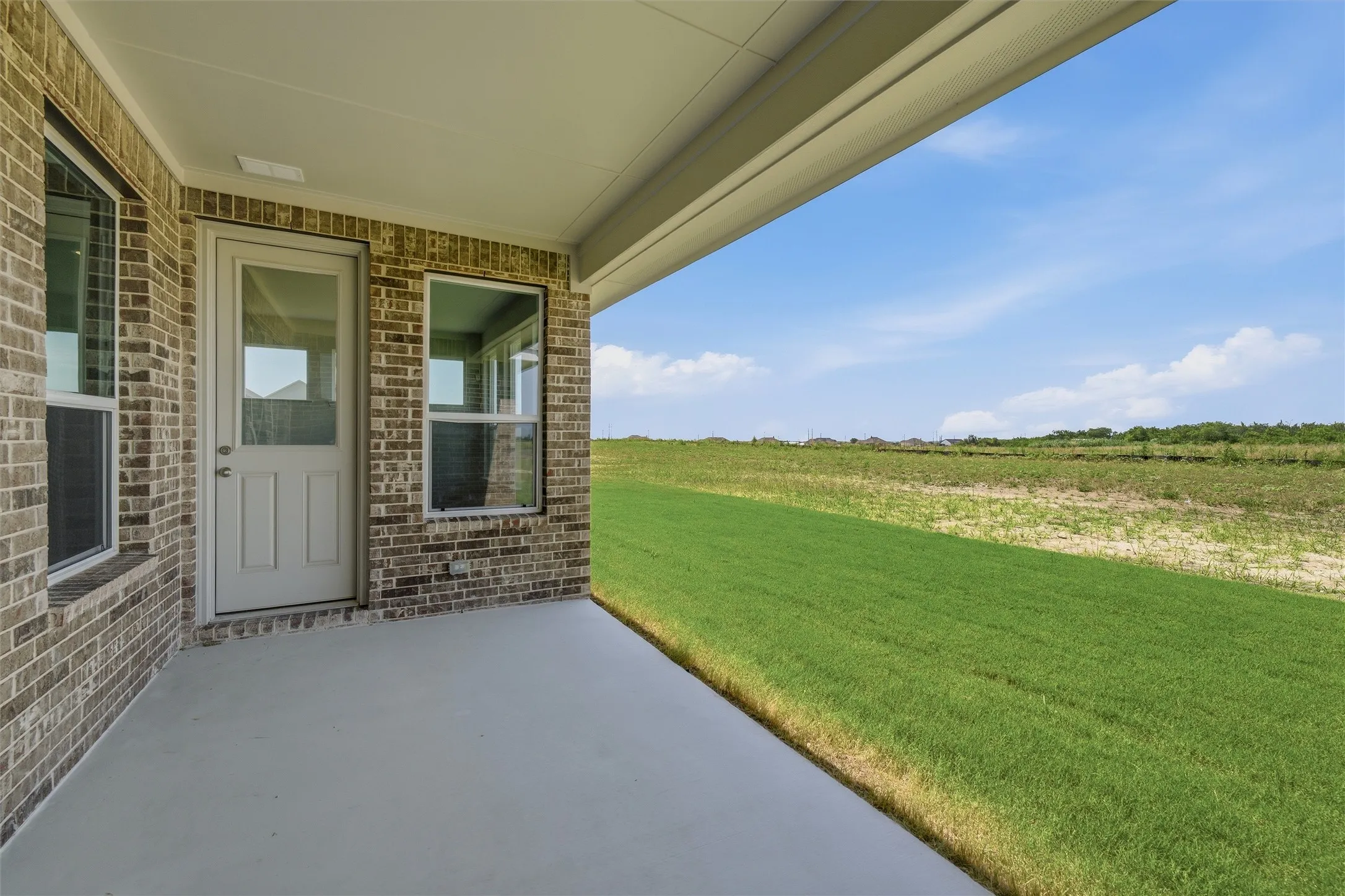 Covered patio with a concrete surface, brick exterior wall, and multiple windows, providing an outdoor living space beside an expanse of green grass