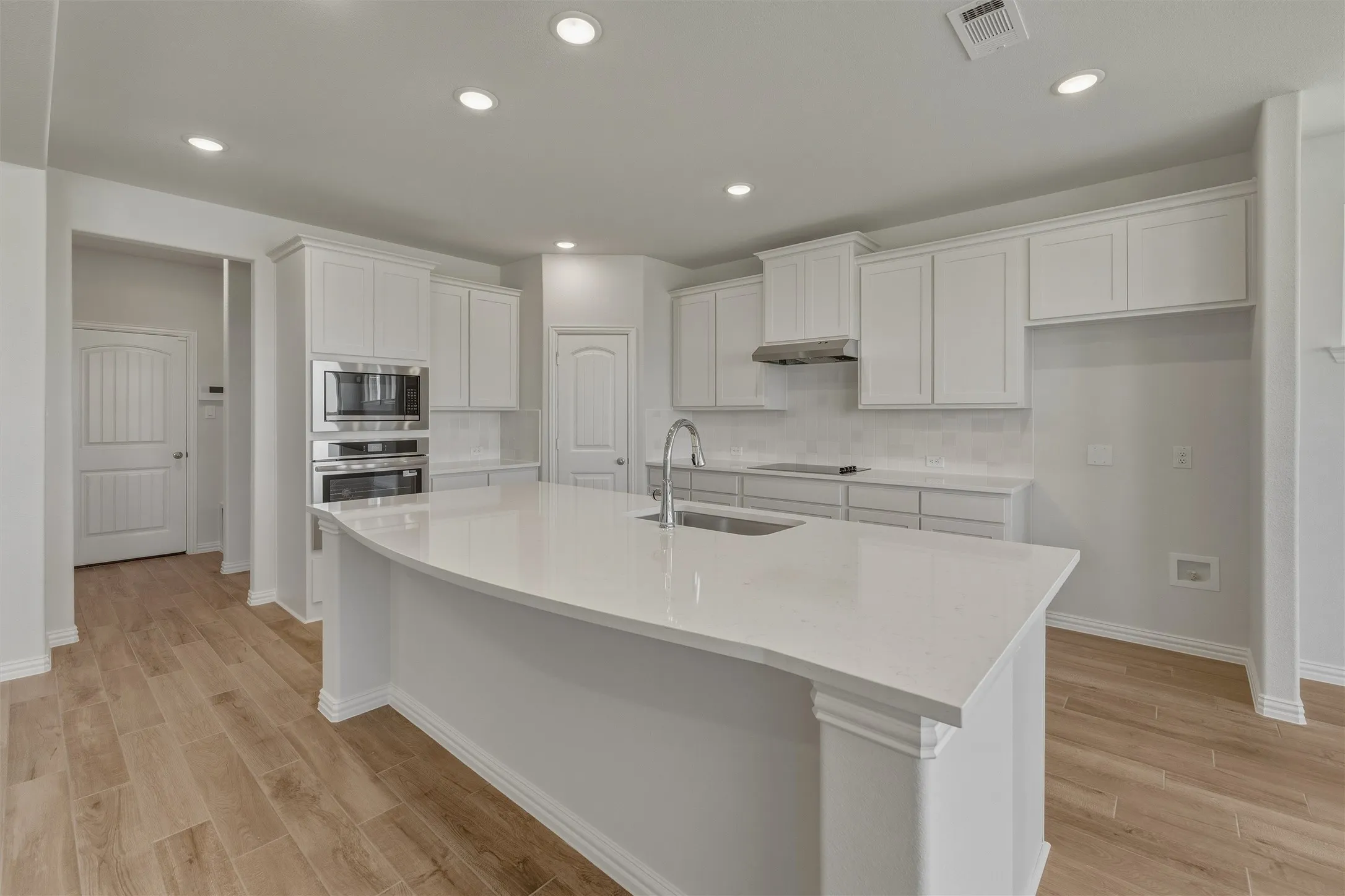Expansive kitchen featuring a large central island with an undermount sink, white shaker cabinetry, stainless steel built-in appliances, a tiled backsplash, and wood-finish flooring