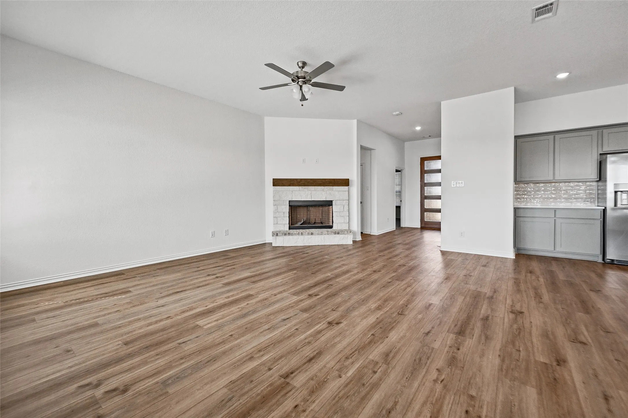 Wood-finish flooring throughout, fireplace with a white brick surround, wood mantel, ceiling fan, and gray kitchen cabinetry with a light-colored backsplash