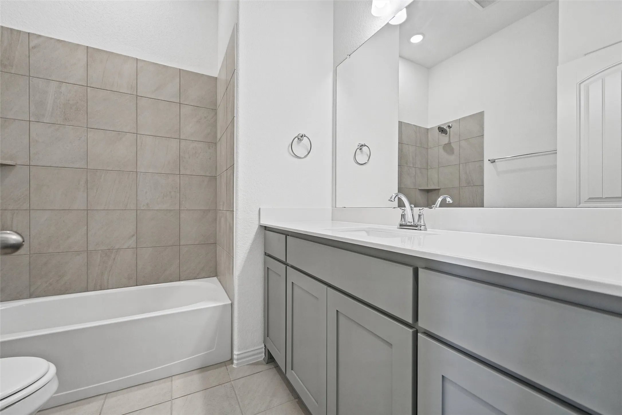 Neutral-toned tile flooring, light gray vanity cabinetry, white countertop, chrome faucet, and a large mirror