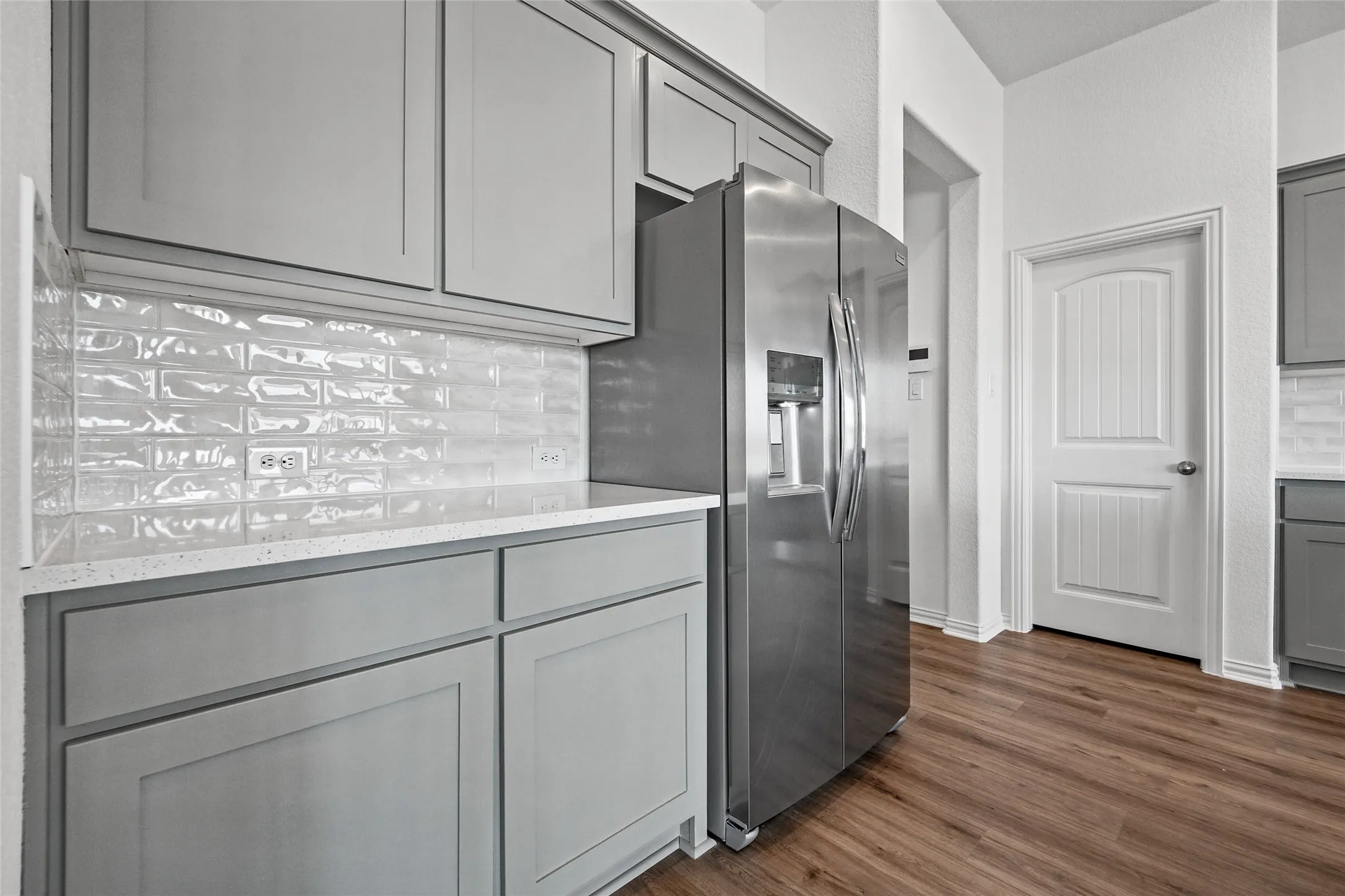Gray shaker-style cabinetry with crown molding, light-toned subway tile backsplash, white flecked countertops, wood-finish flooring, and a stainless steel refrigerator