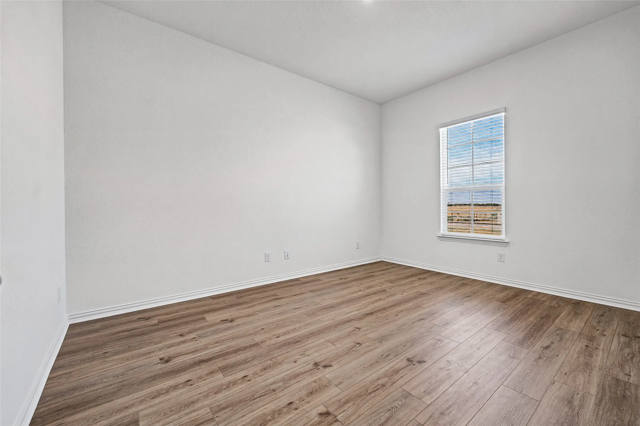 Wood-finish flooring with white baseboards, a single window with blinds, and white painted walls