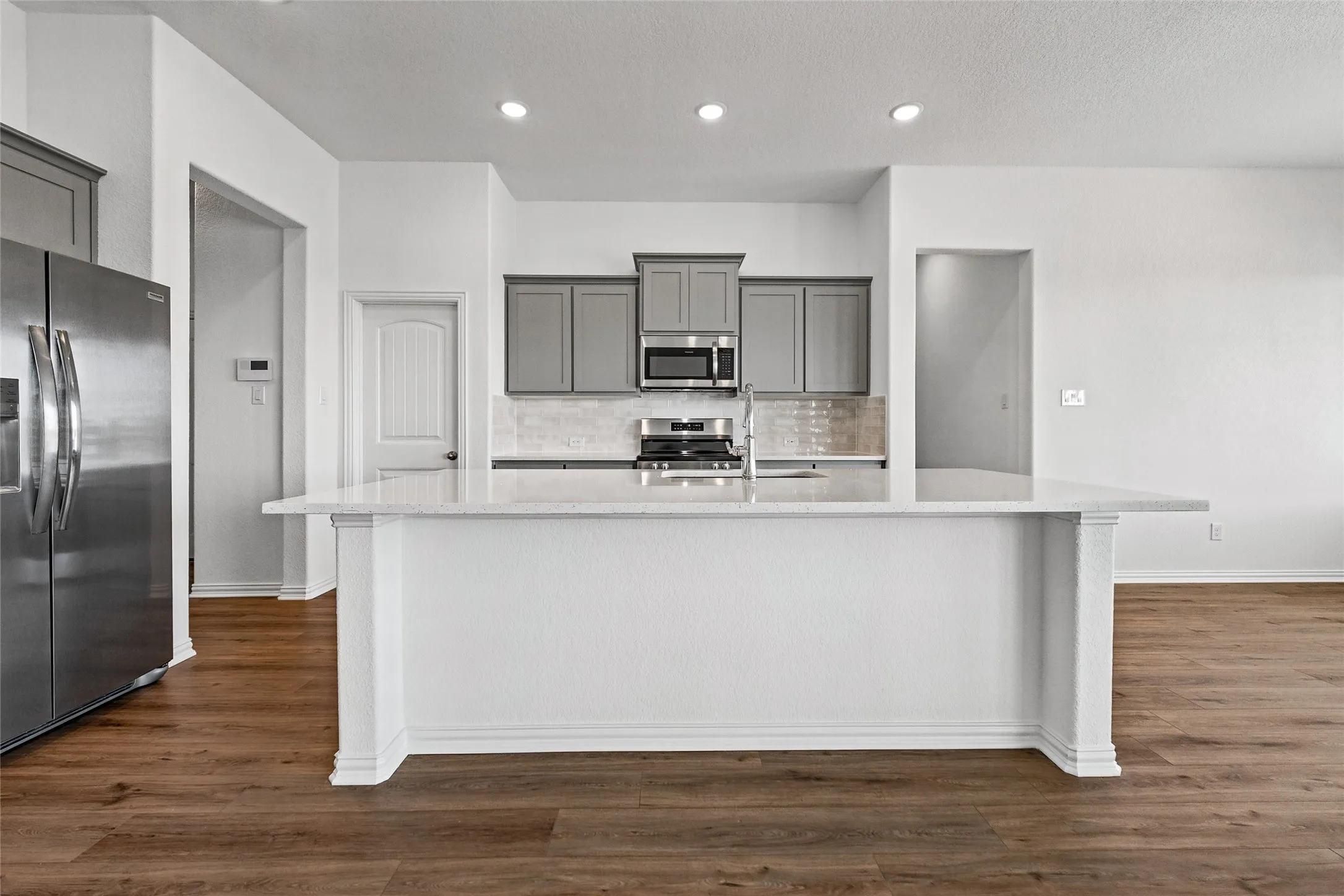 Kitchen featuring an expansive island with a light-toned countertop, wood-finish flooring, gray shaker-style cabinetry, and stainless steel appliances
