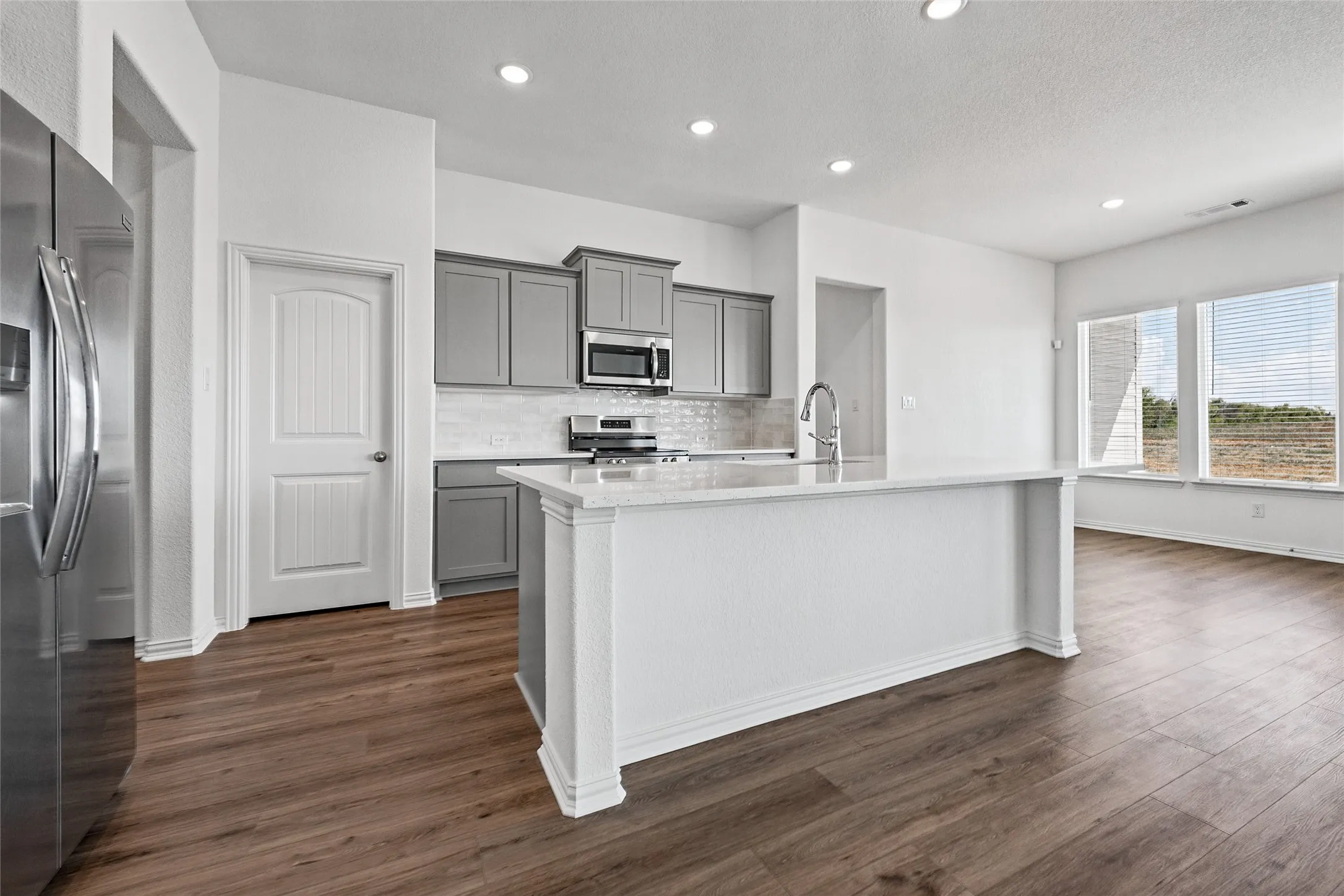 Kitchen featuring wood-finish flooring, gray cabinetry, a white tile backsplash, a stainless steel microwave, and a kitchen island with a white countertop
