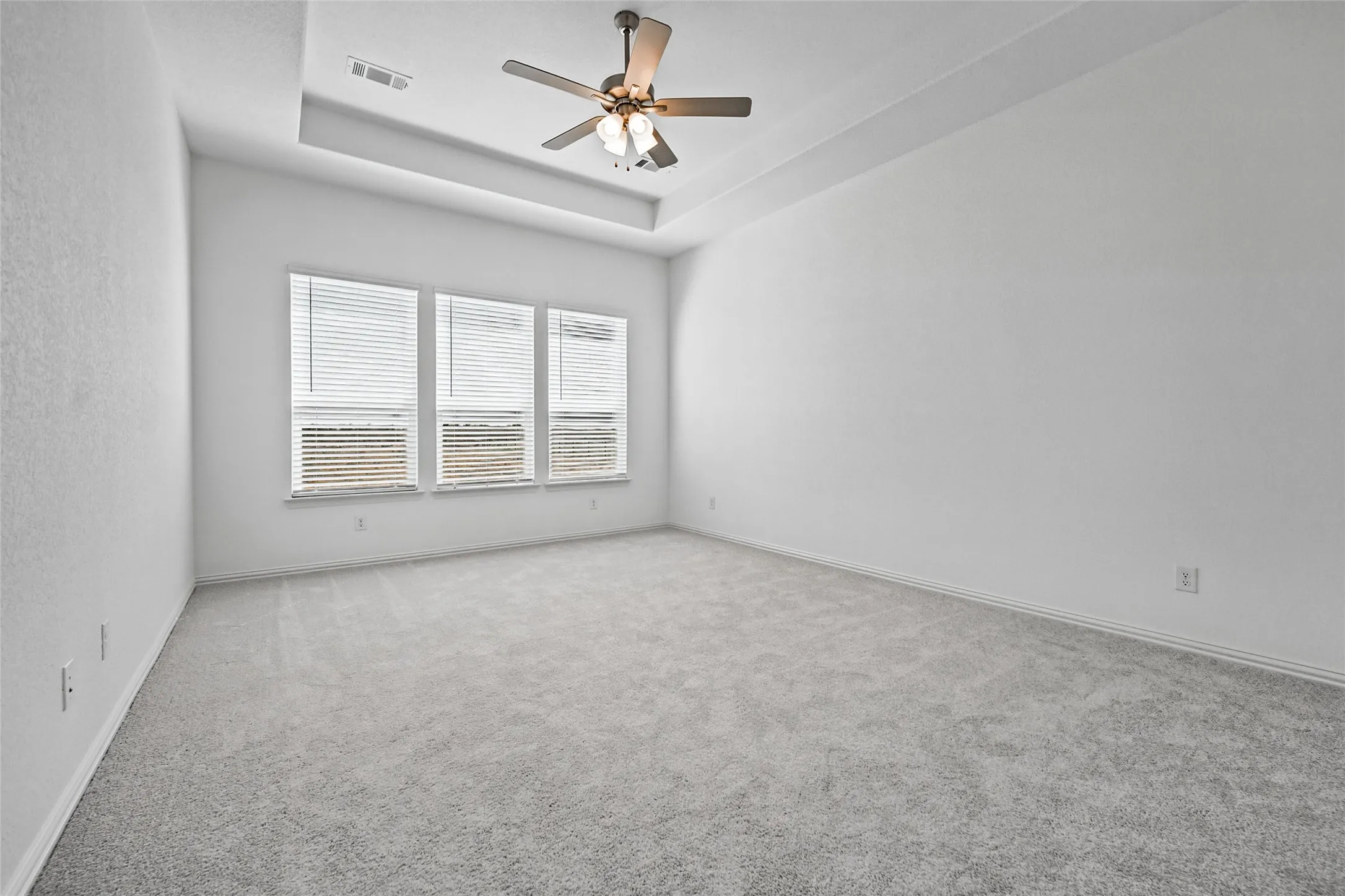 Recessed tray ceiling with a ceiling fan, three windows with blinds, and light-colored carpet
