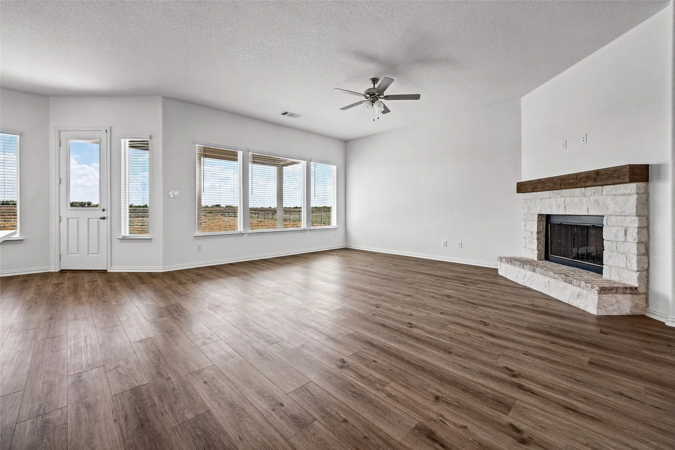 Spacious room featuring wood-finish flooring, a stone fireplace with a wood-beam mantle, multiple windows with blinds, a ceiling fan, and a white door with a window insert