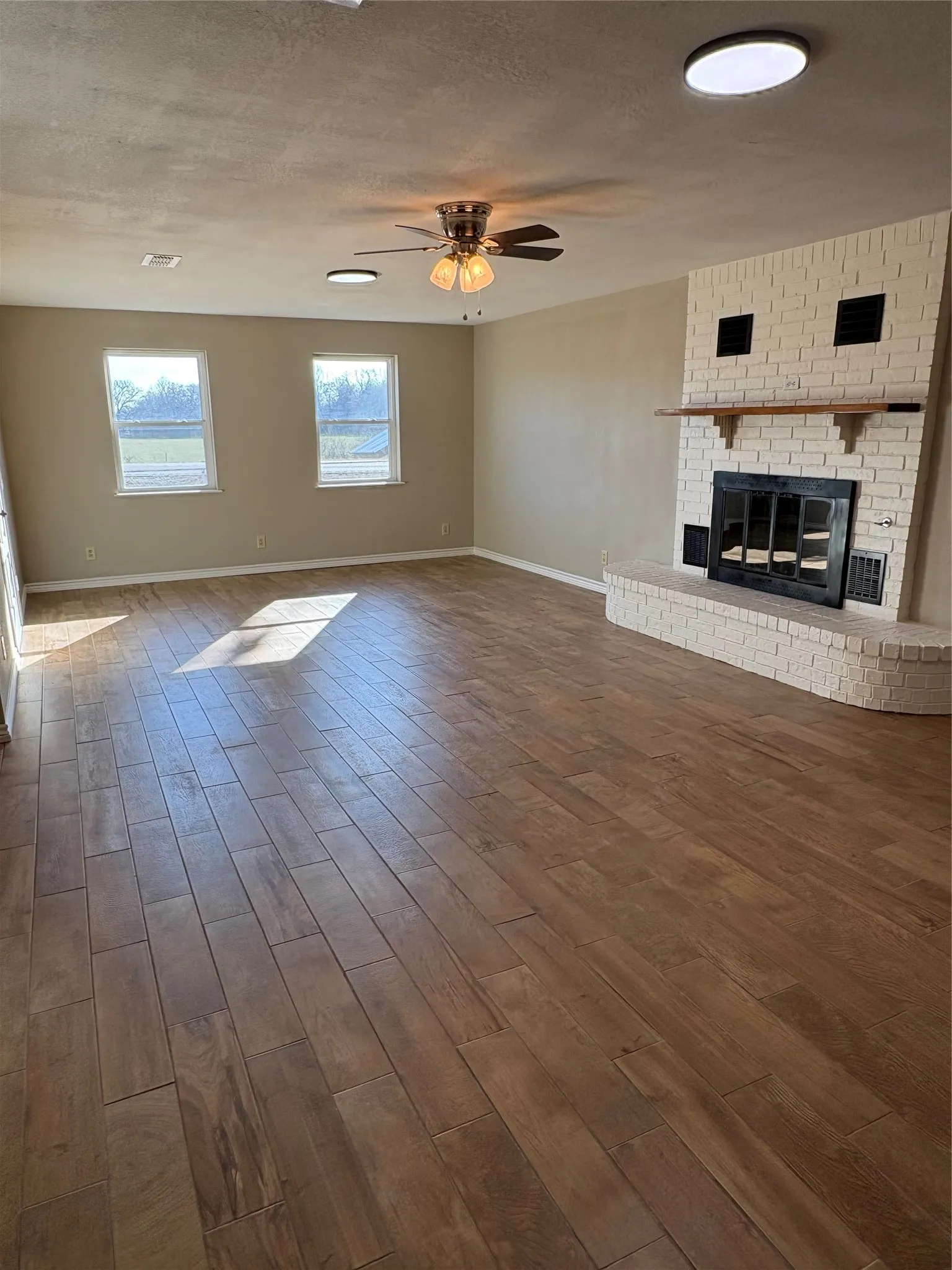 Unfurnished living room with a brick fireplace, dark wood-style flooring, a ceiling fan, and good exterior lighting.
