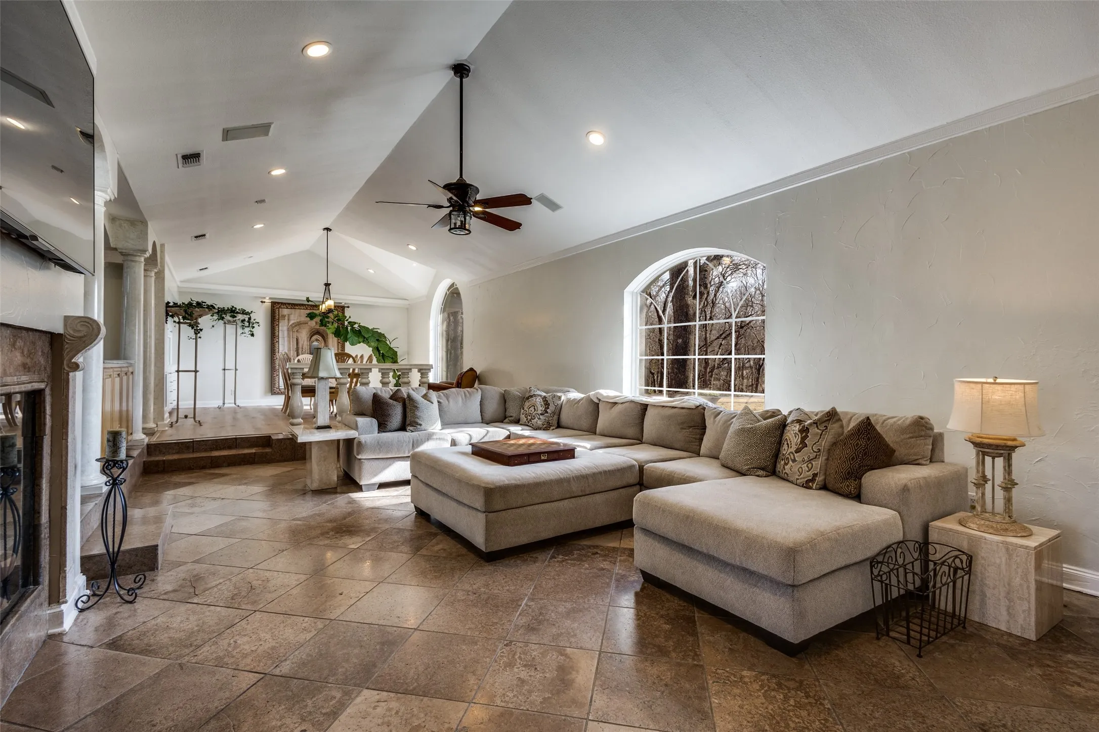 Split-level living room with vaulted ceiling, walnut travertine flooring, and arched window with wooded views. Step up to adjoining dining area framed by marble columns for an open yet defined layout. Ideal for gathering and entertaining with multiple seating zones.