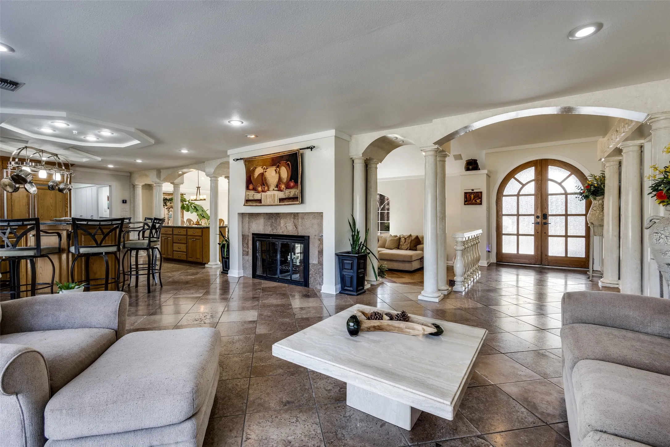 Arched Mahogany double doors with divided light windows serve as a striking architectural focal point. Marble columns and curved openings frame the entry and living spaces for a classic Tuscan-inspired feel. Walnut travertine flooring ties the areas together for a cohesive look.