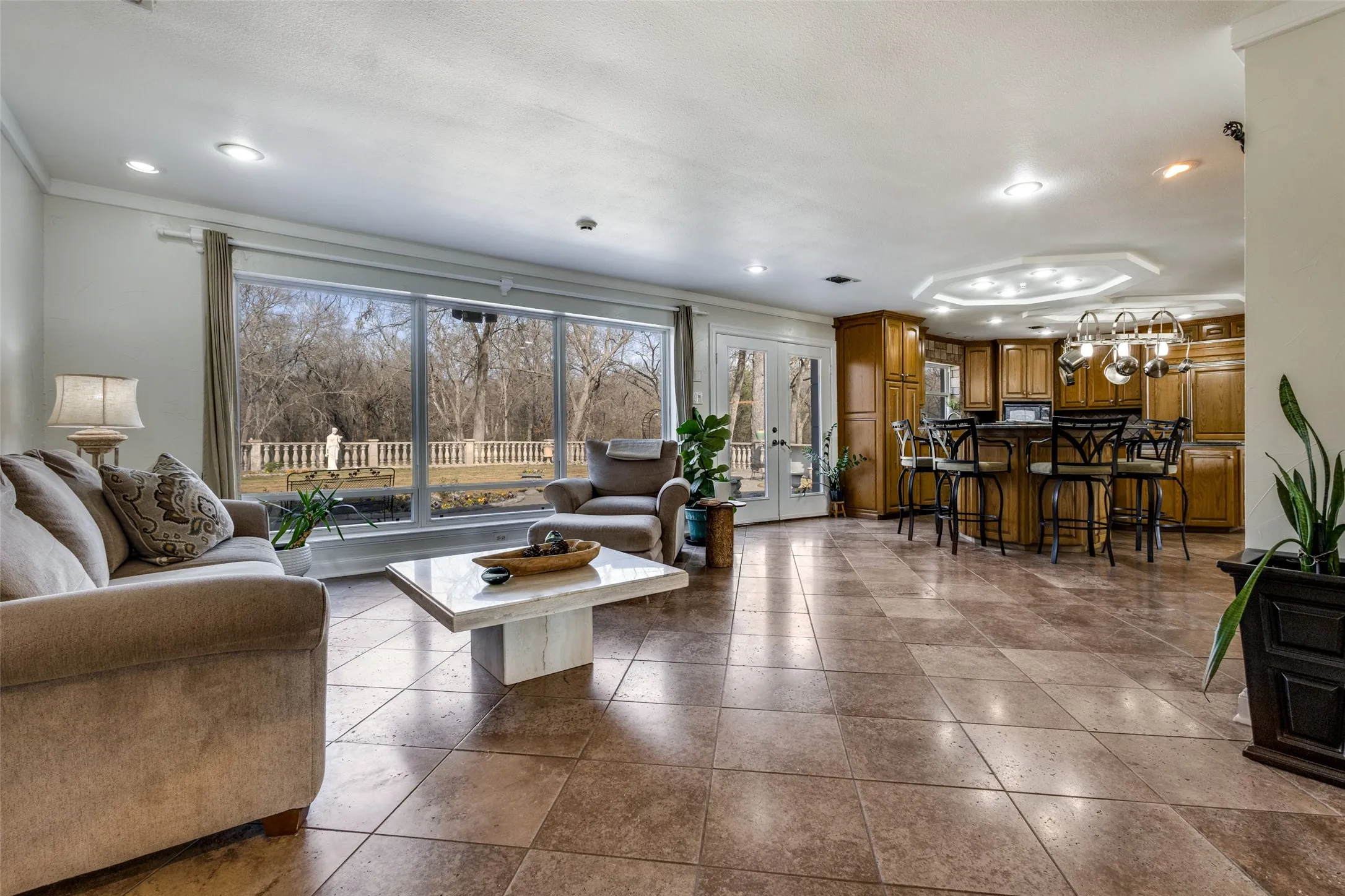 Spacious living and dining area with large picture windows overlooking the Tuscan-style garden and wooded surroundings. Walnut travertine tile continues through the space for a cohesive look. Open layout connects directly to the kitchen and outdoor access for indoor-outdoor living.