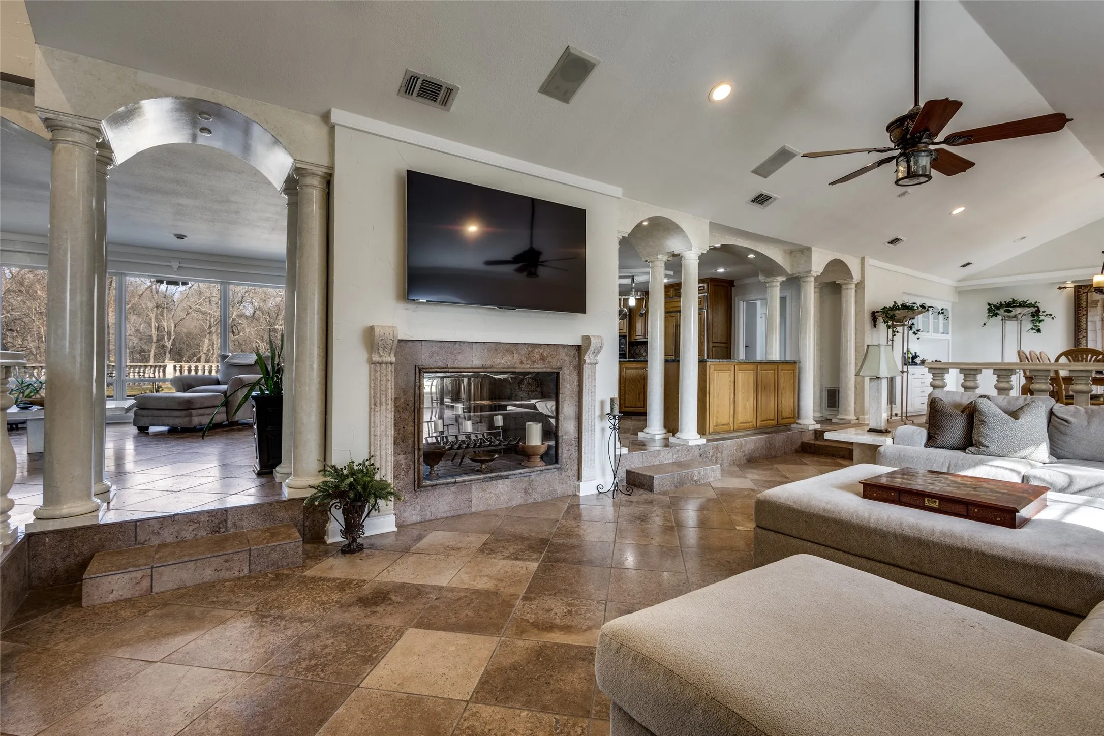 Main living area with walnut travertine flooring, see-through fireplace, and marble column accents. Arched openings connect the adjacent sunroom and kitchen for an open flow. Large rear windows provide natural light and wooded property views.