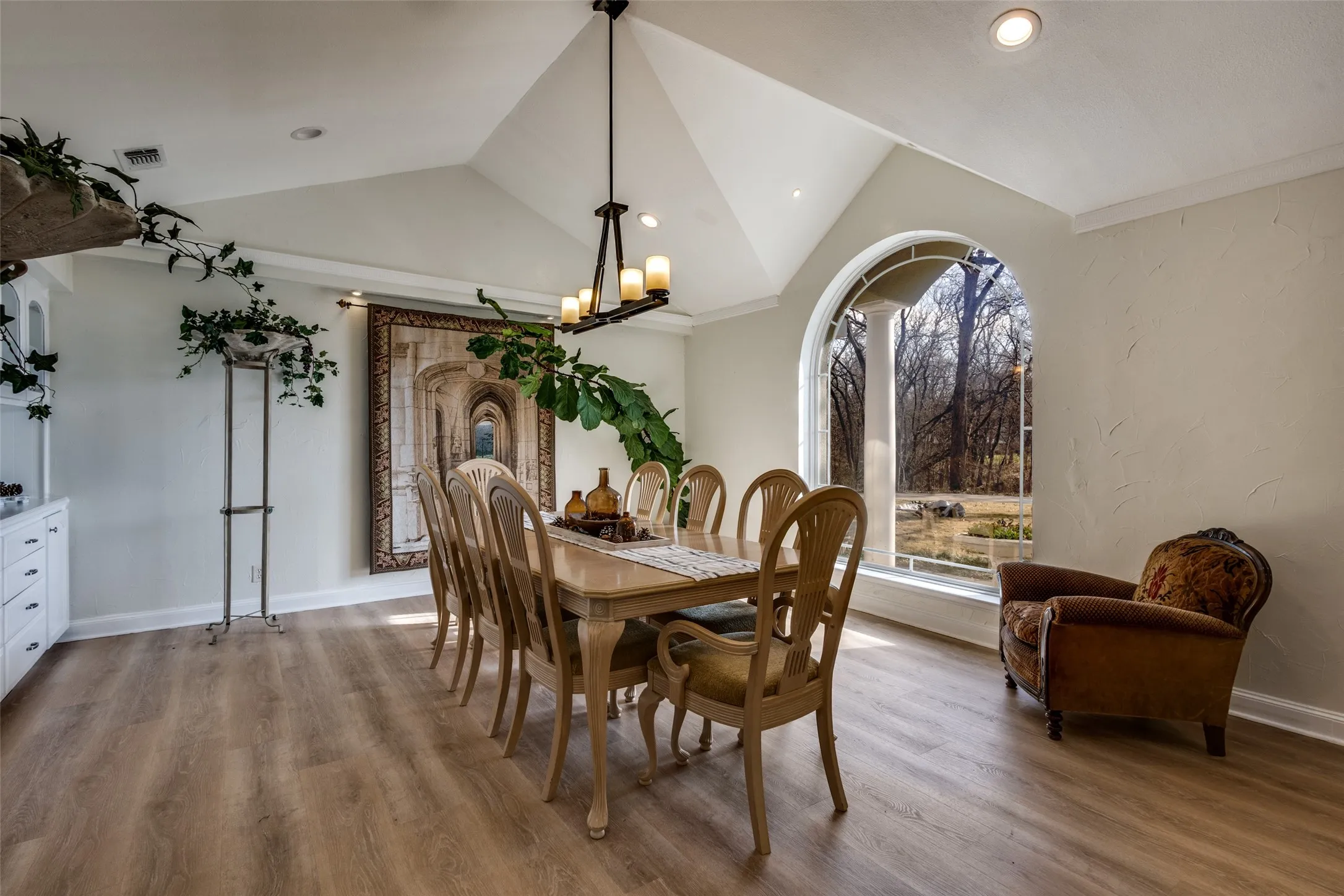 Dining room with vaulted ceiling, built-in cabinetry for china and serving storage, and a large arched window overlooking wooded views. Spacious layout accommodates a full dining table for entertaining. Natural light highlights the open, inviting feel of the space.