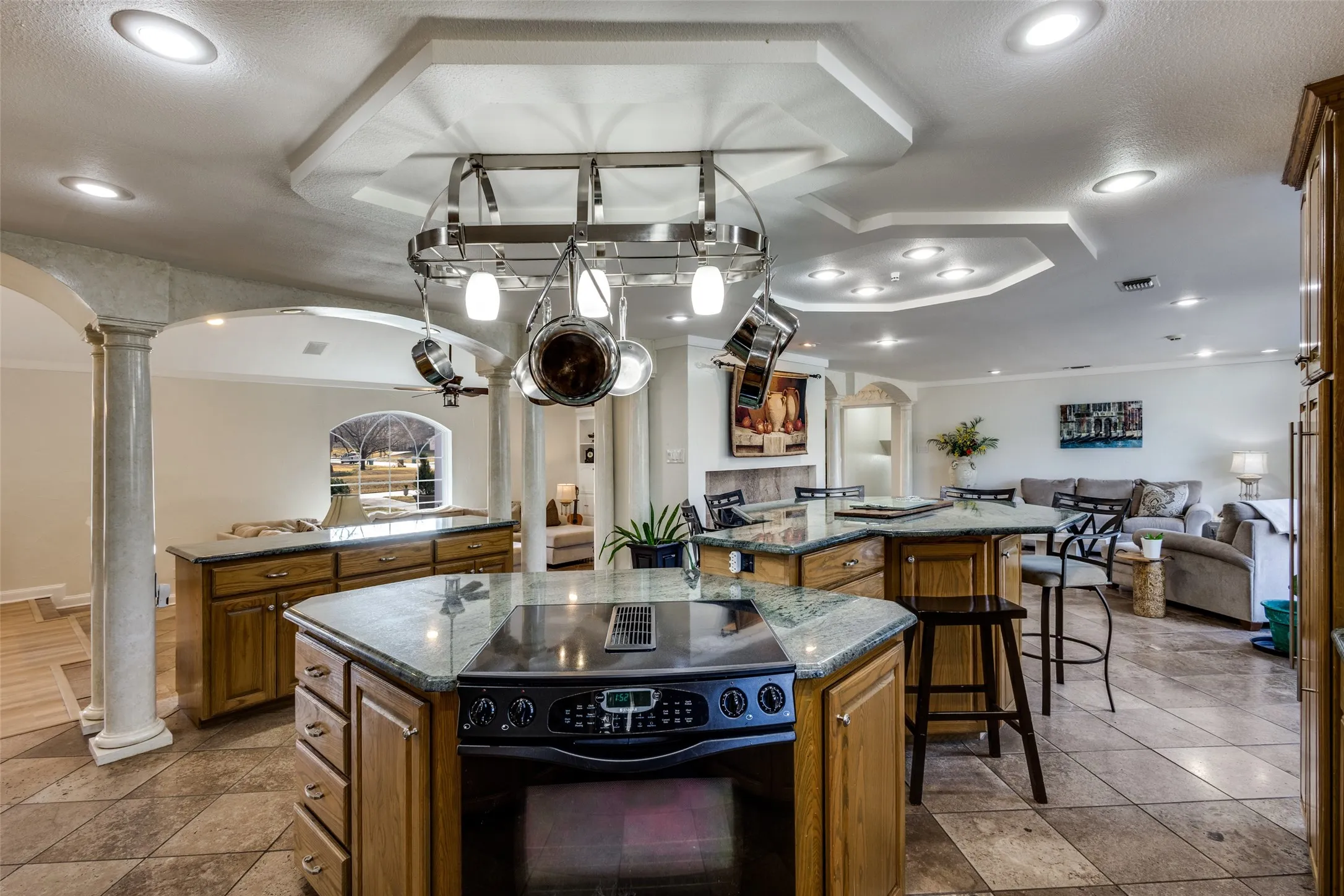 Center island with electric cooktop and overhead pot rack anchors the open kitchen layout. Granite counters, warm wood cabinetry, and columned openings blend with the adjacent living and dining areas. Multi-level ceilings with recessed lighting add dimension and character to the space.